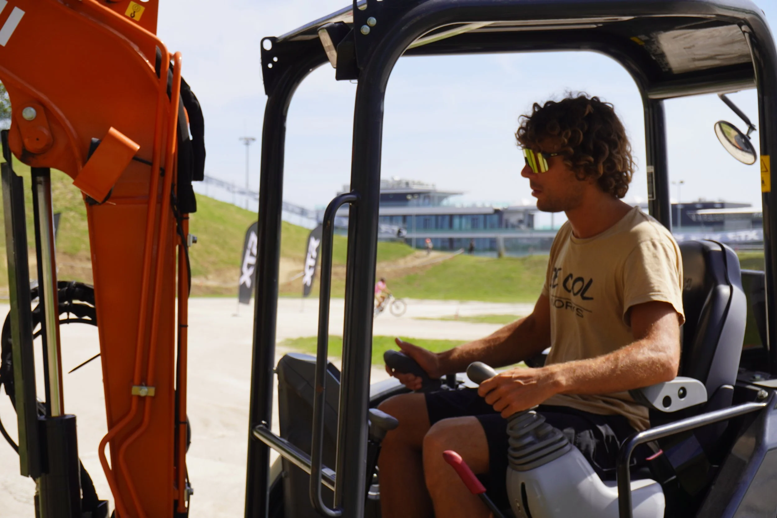 Uomo seduto in un escavatore senza cabina, indossa occhiali da sole e tiene le leve di controllo, con un'area di campagna e edifici sullo sfondo.