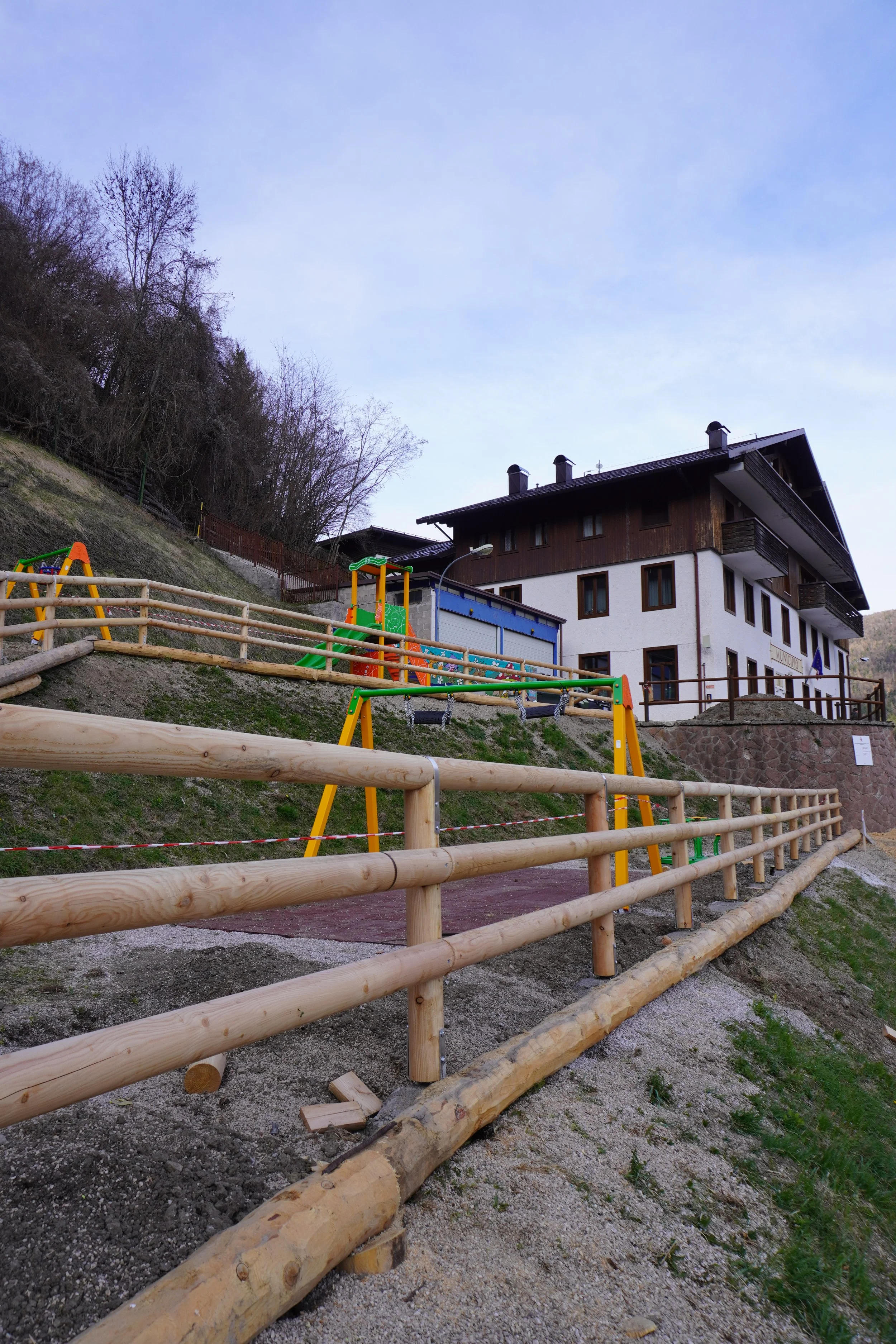Parco giochi con altalene in legno di fronte a un edificio in stile alpino, situato su una collina con alberi spogli circostanti.