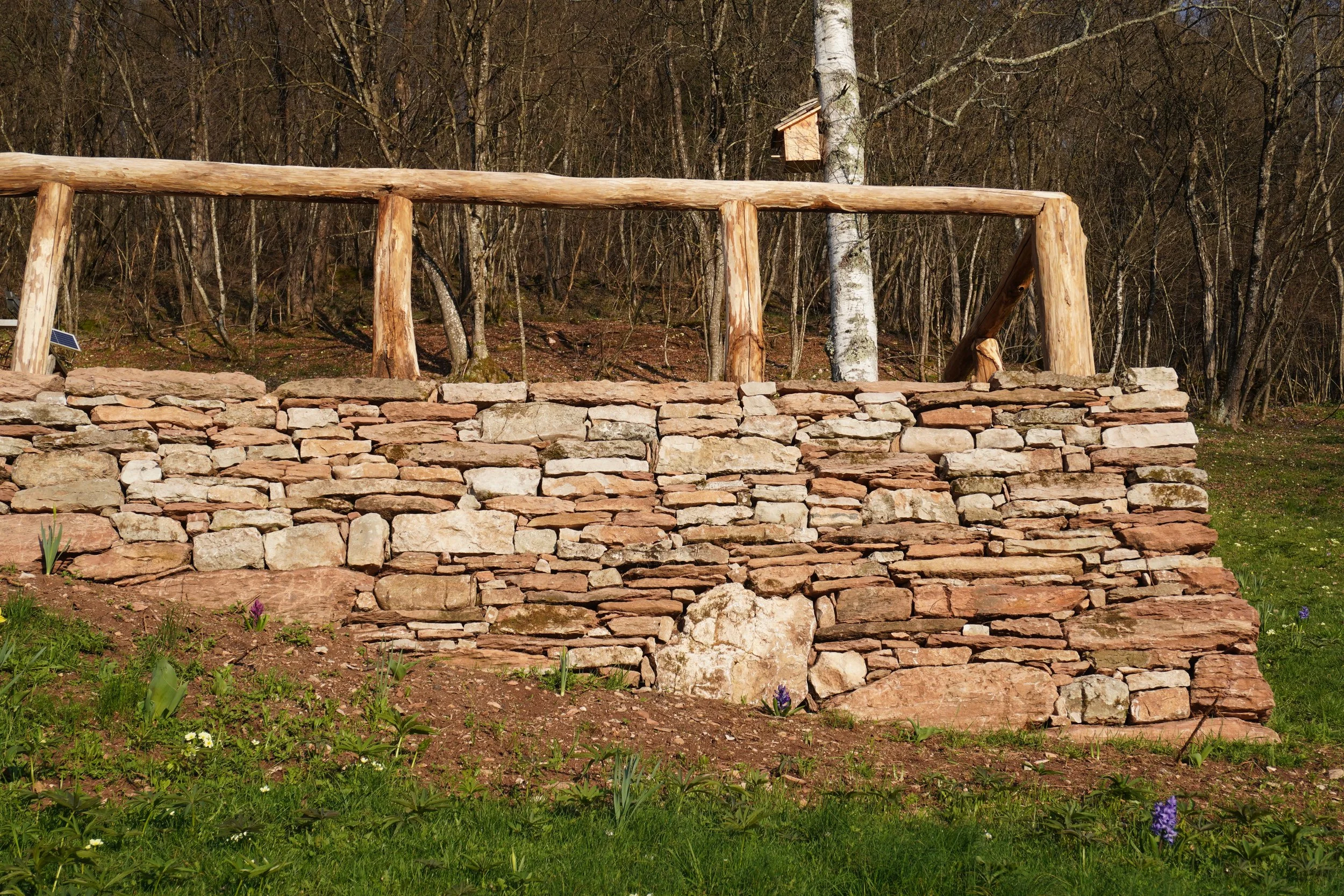 Muretto in pietra naturale con ringhiera in legno, natura circostante con alberi e fiori di primavera.