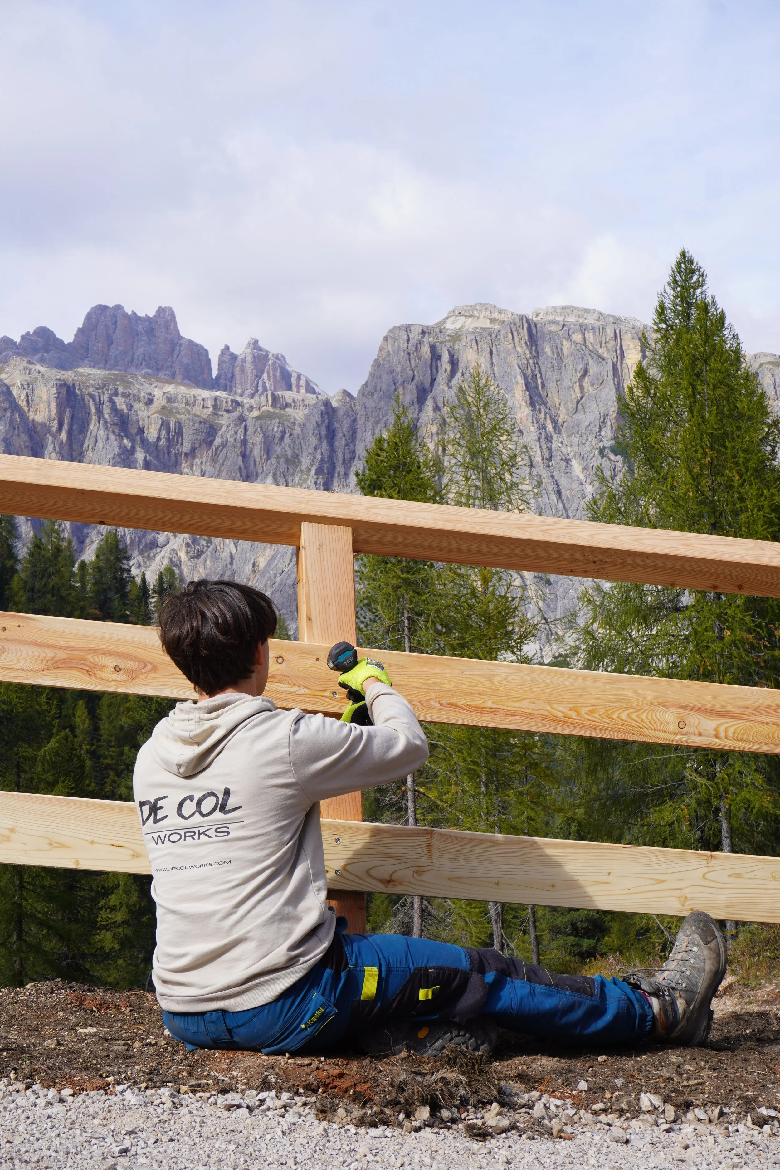 Un giovane sta installando una recinzione di legno in un'area di montagna con alte pareti di roccia e alberi verdi sullo sfondo.