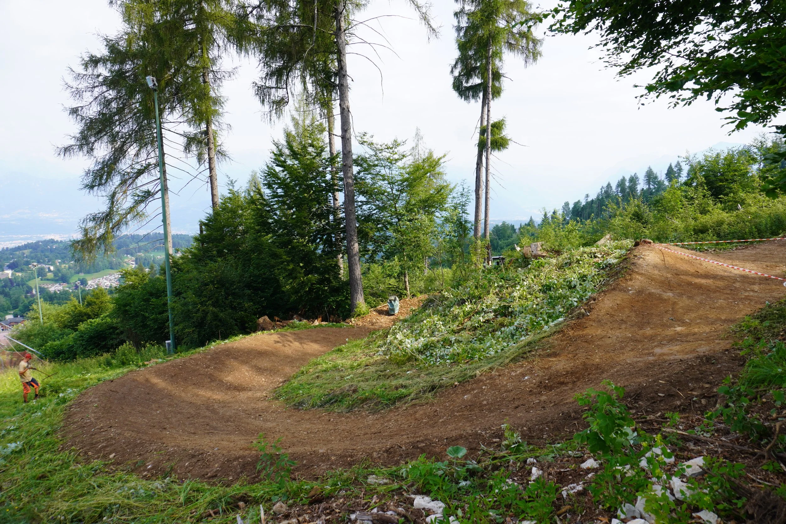 Sentiero di mountain bike in un bosco, con alberi alti e fogliame verde, e un paesaggio di colline e città sullo sfondo.