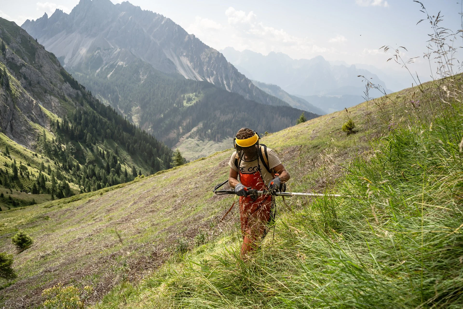 Uomo che utilizza un tosaerba a benzina su un prato in una zona montuosa con montagne e boschi sullo sfondo.