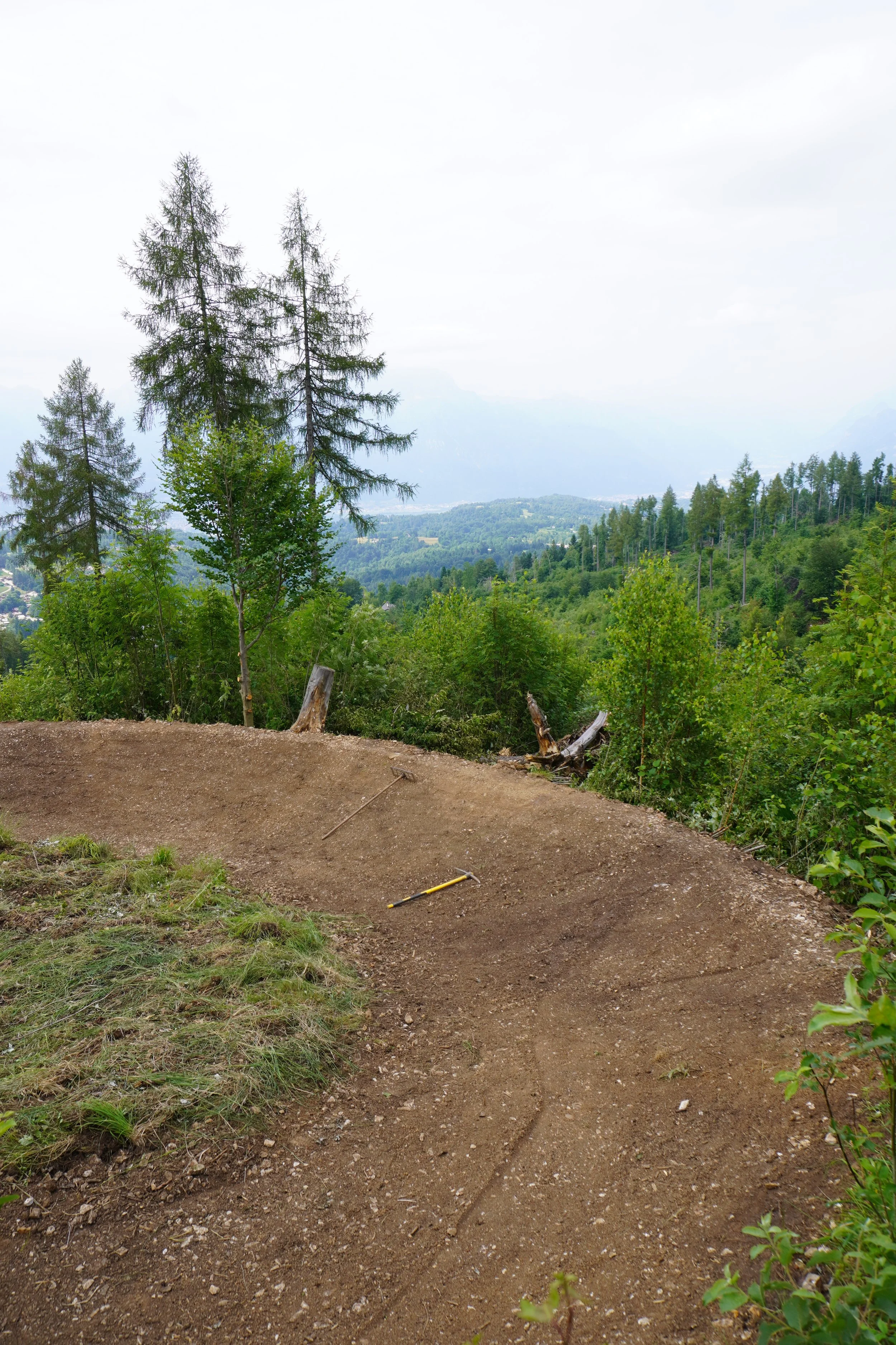Sentiero di montagna in fase di costruzione o manutenzione, con attrezzi sul terreno, circondato da alberi e una vista panoramica sulla valle e le montagne lontane.
