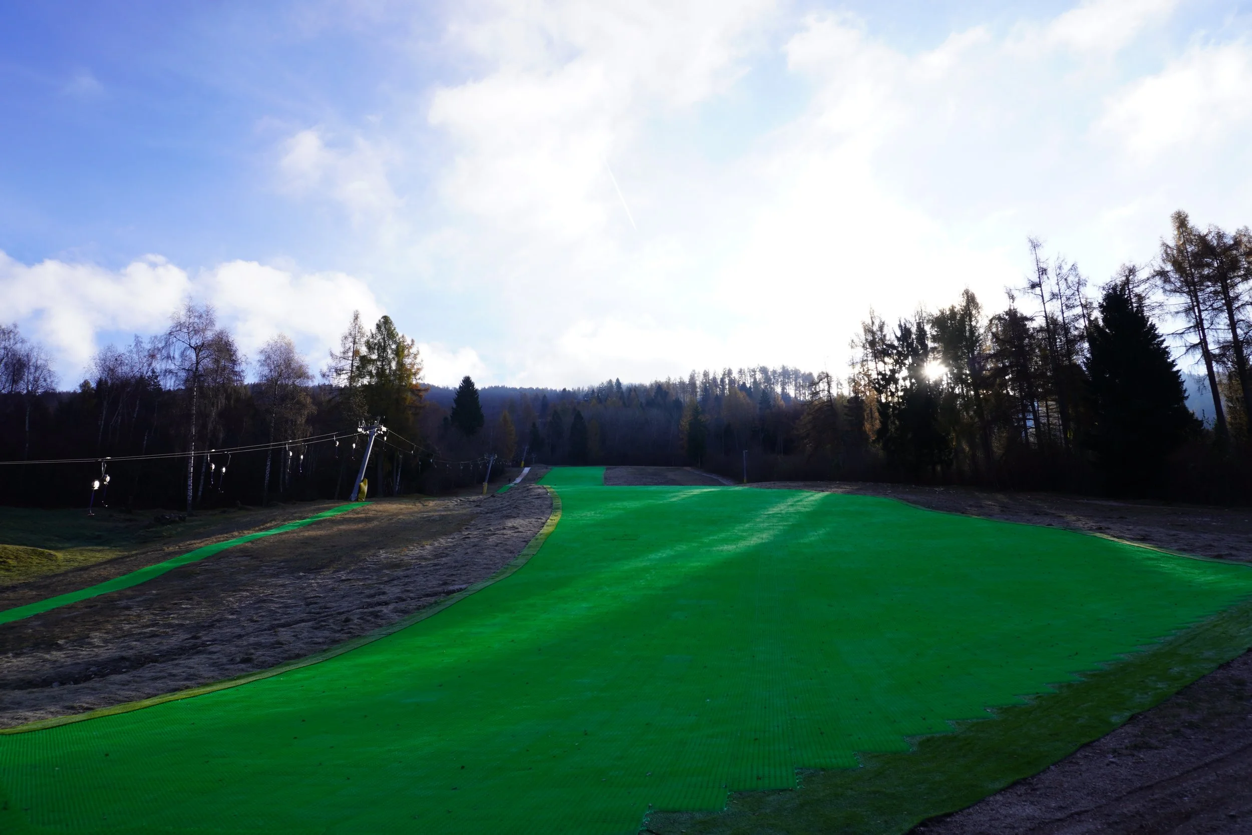 Pista da sci in una località montana, con una superficie coperta di manto verde e contorni naturali di alberi e cielo nuvoloso.