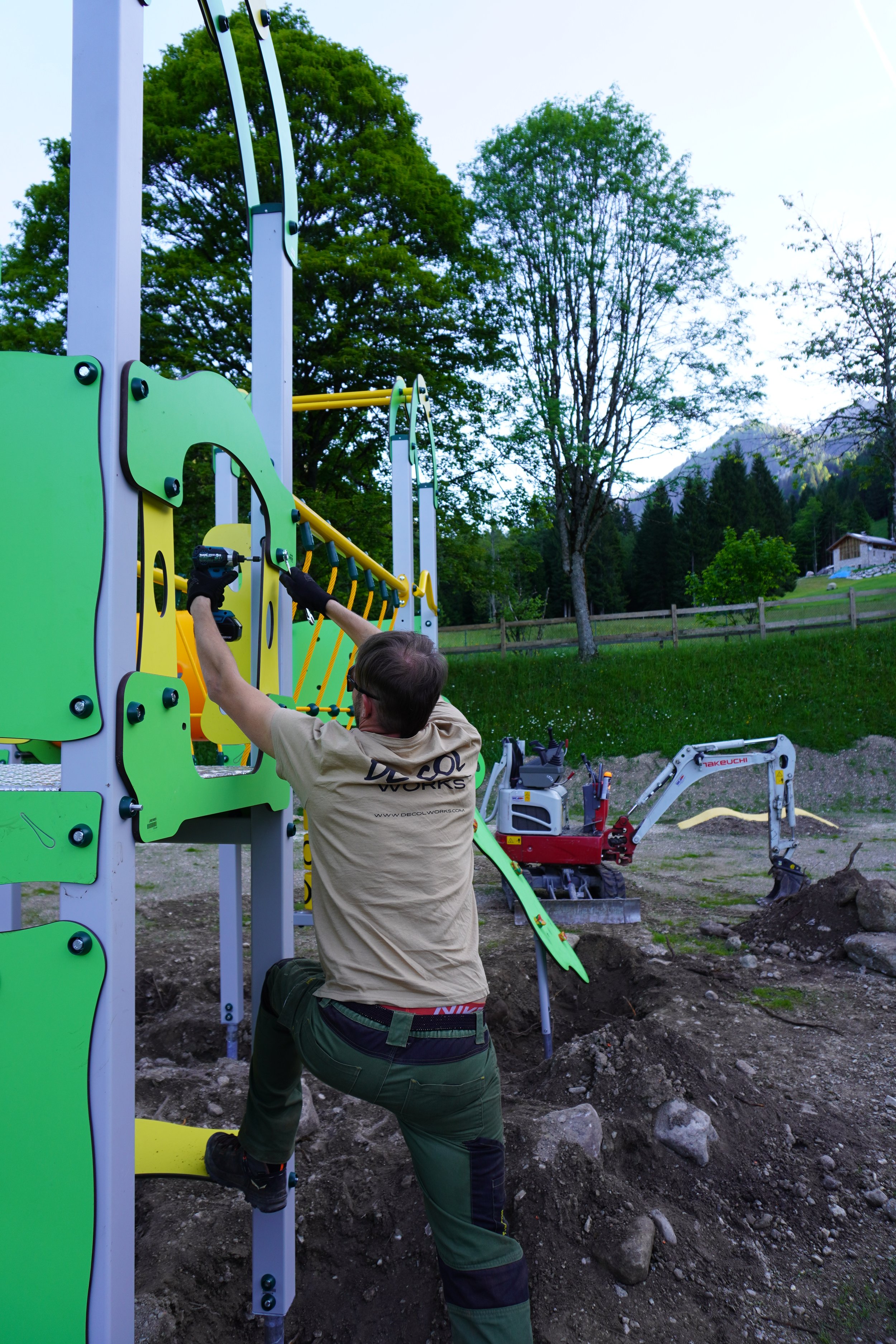 Lavoratore installa un'area giochi con attrezzature verdi e gialle in un parco all'aperto, con alberi e una casa sullo sfondo.