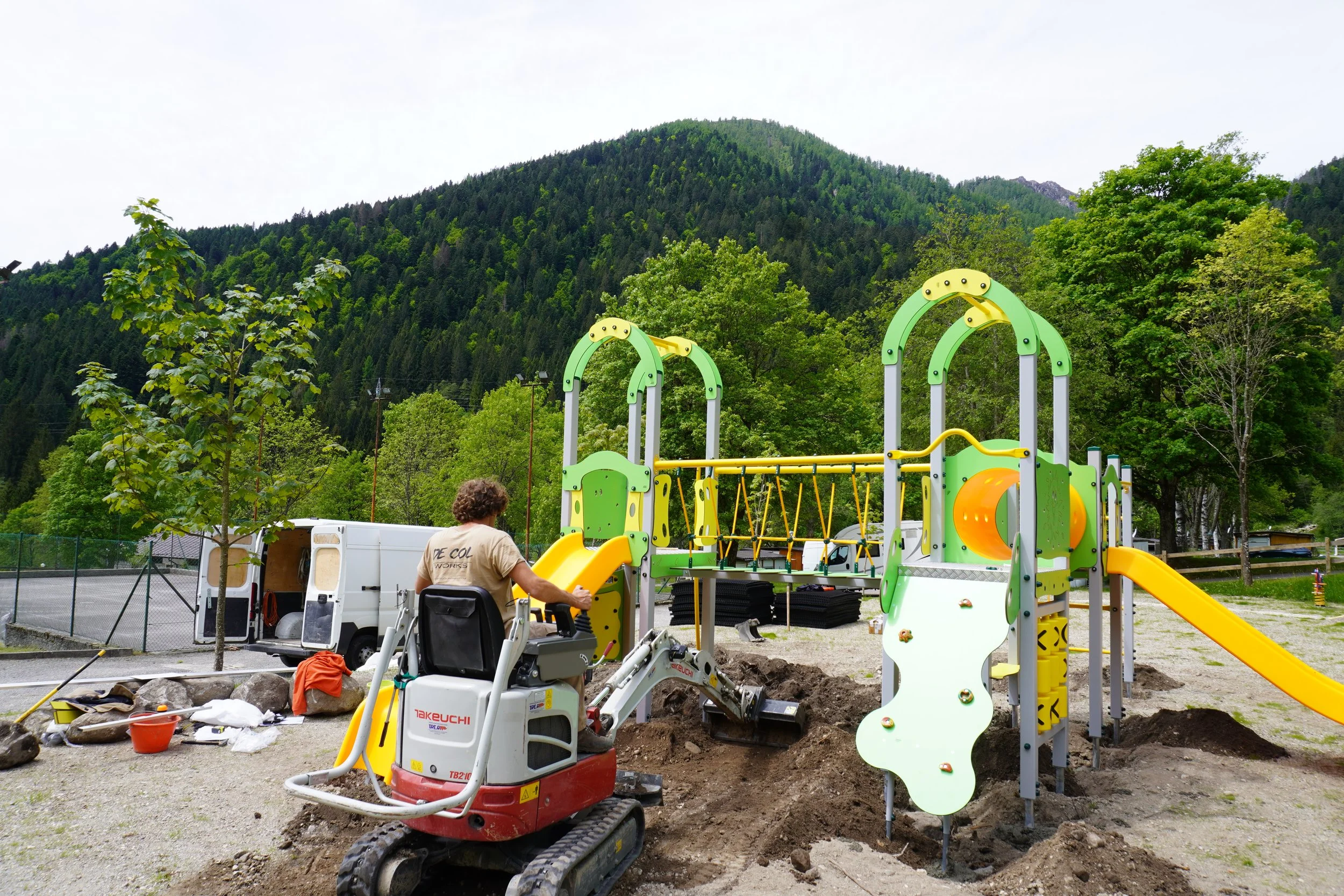 Lavoratore su una mini escavatrice mentre lavora in un cantiere di un parco giochi in fase di costruzione, con sottoe e attrezzi intorno, con un'area verde e montagne sullo sfondo.