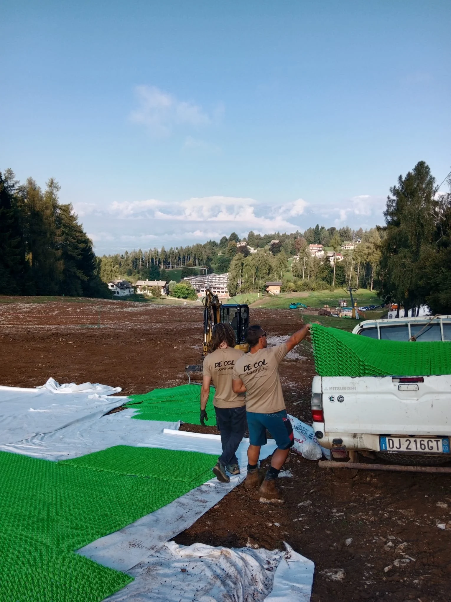 Due lavoratori stanno posando pannelli verdi sul terreno, con una camionetta bianca dietro. Sfondo di un paesaggio collinare e cielo azzurro con nuvole.