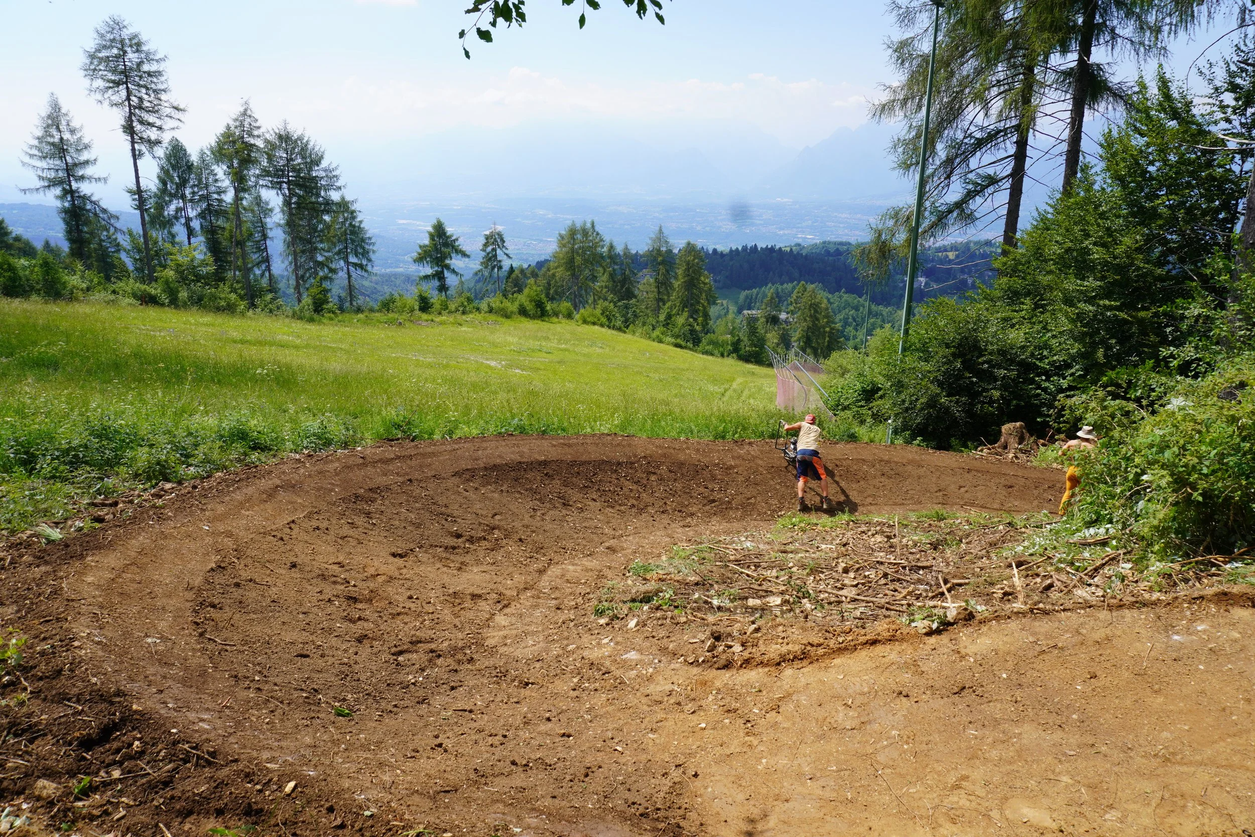 Due bambini giocano sul terreno scavato in una zona collinare circondata da alberi e vegetazione, con un paesaggio montano sullo sfondo.