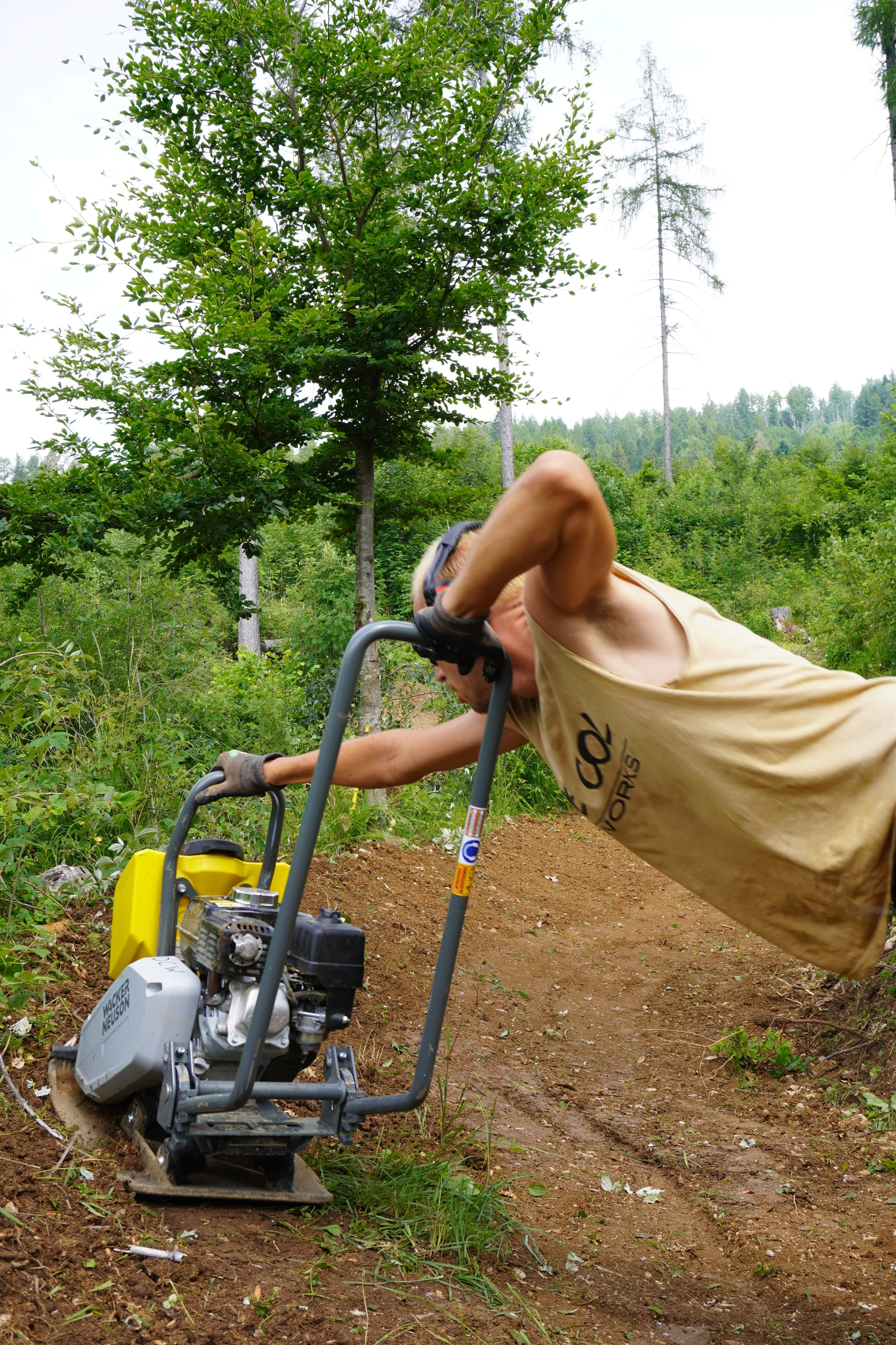 Uomo che lavora con una macchina trapanatrice a motore su un sentiero nel bosco, con alberi verdi e tracce di terra.