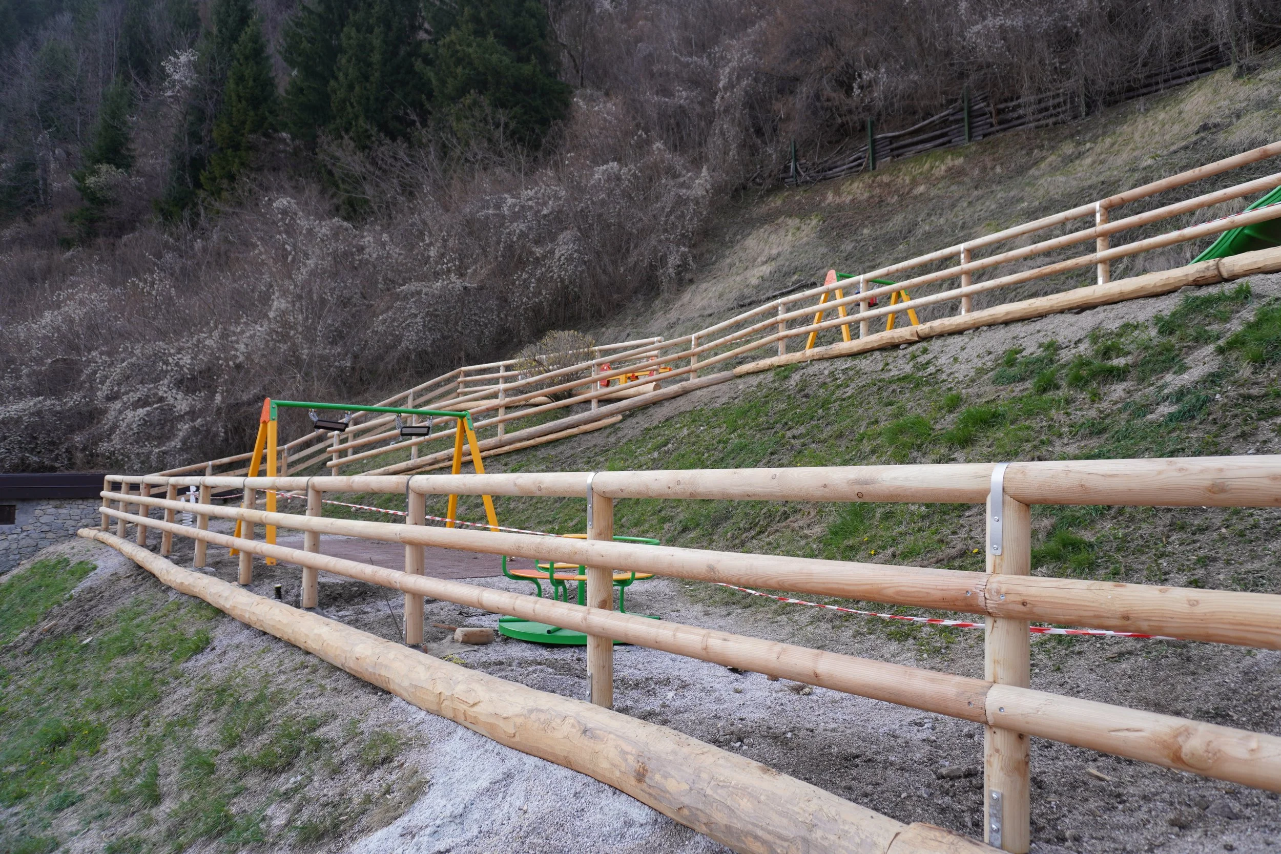 Barriera in legno e giochi per bambini installati su un pendio naturale, con alberi e terreno erboso sullo sfondo.