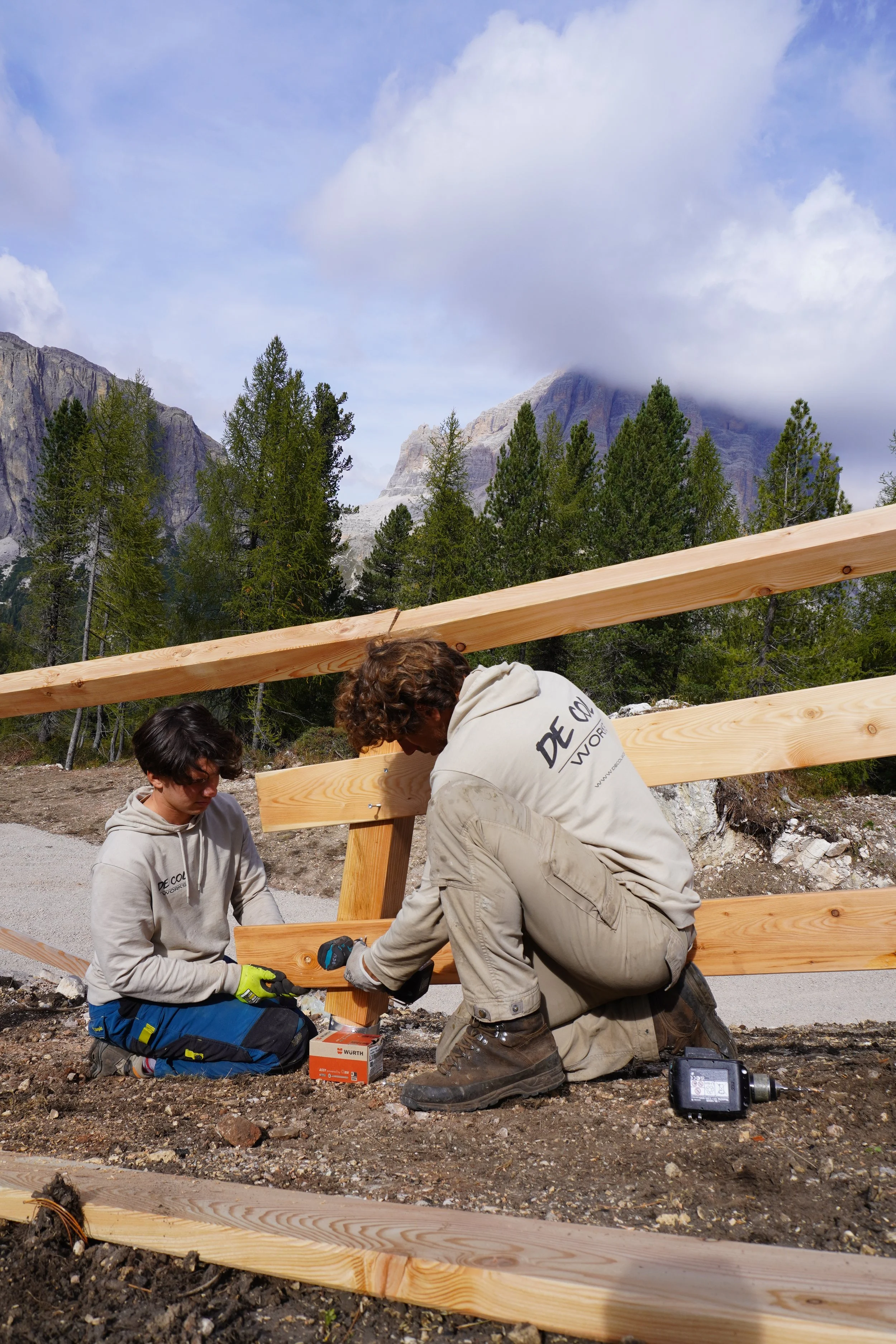 Due persone stanno costruendo una recinzione di legno in un paesaggio montano con alberi e montagne in lontananza, alcuni coperti di nuvole.