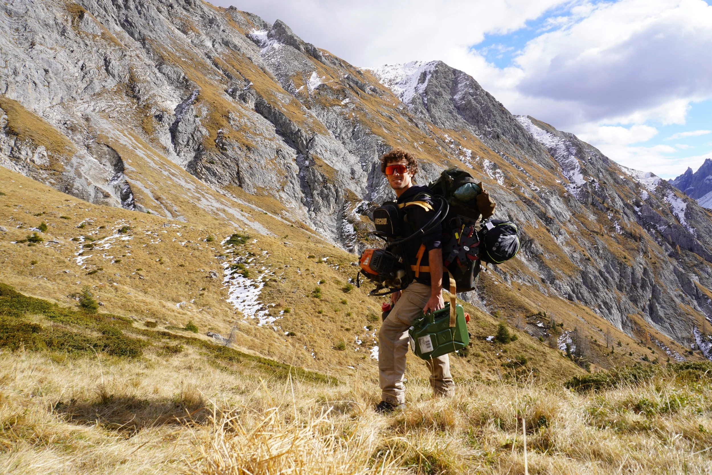 Escursionista con zaino e attrezzatura in un paesaggio montano con erba secca e scogliere con neve