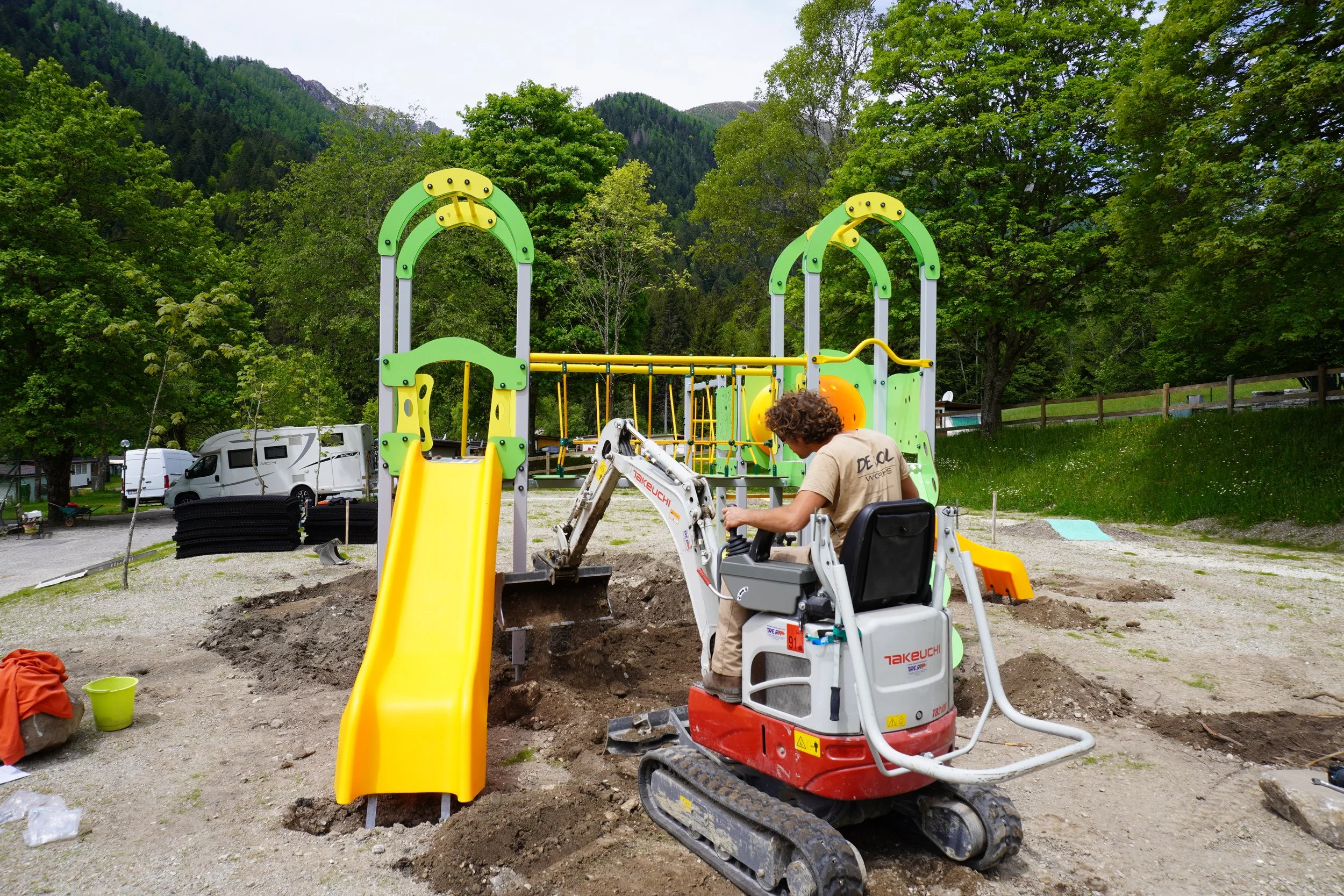 Un uomo sta usando una piccola escavatrice per lavorare sulla costruzione di un'altalena in un parco giochi, con un paesaggio di montagna e alberi sullo sfondo.