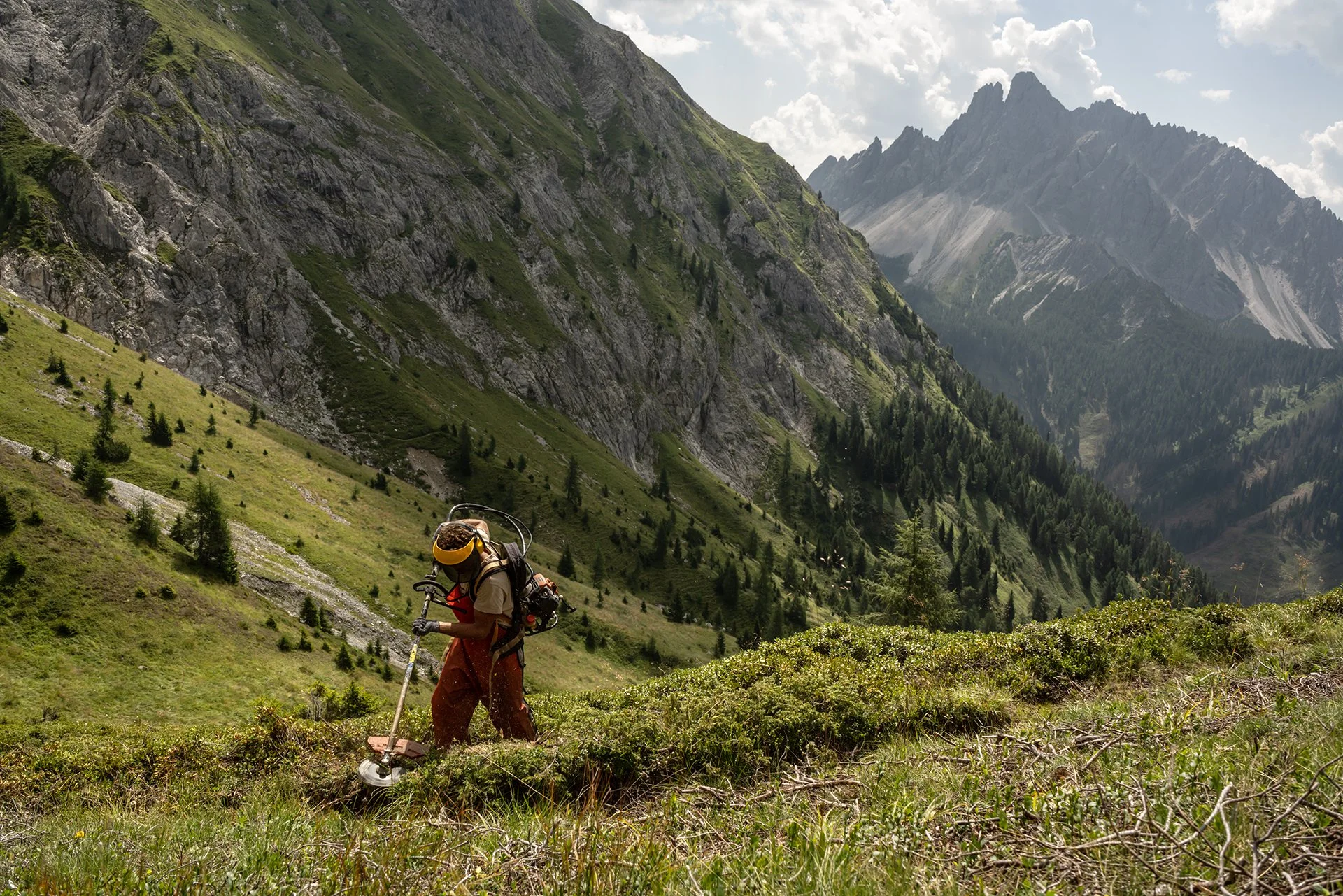 Escursionista con attrezzi da trekking che lavora sulla terra in un paesaggio montano con pendii verdi, alberi e montagne rocciose in lontananza.