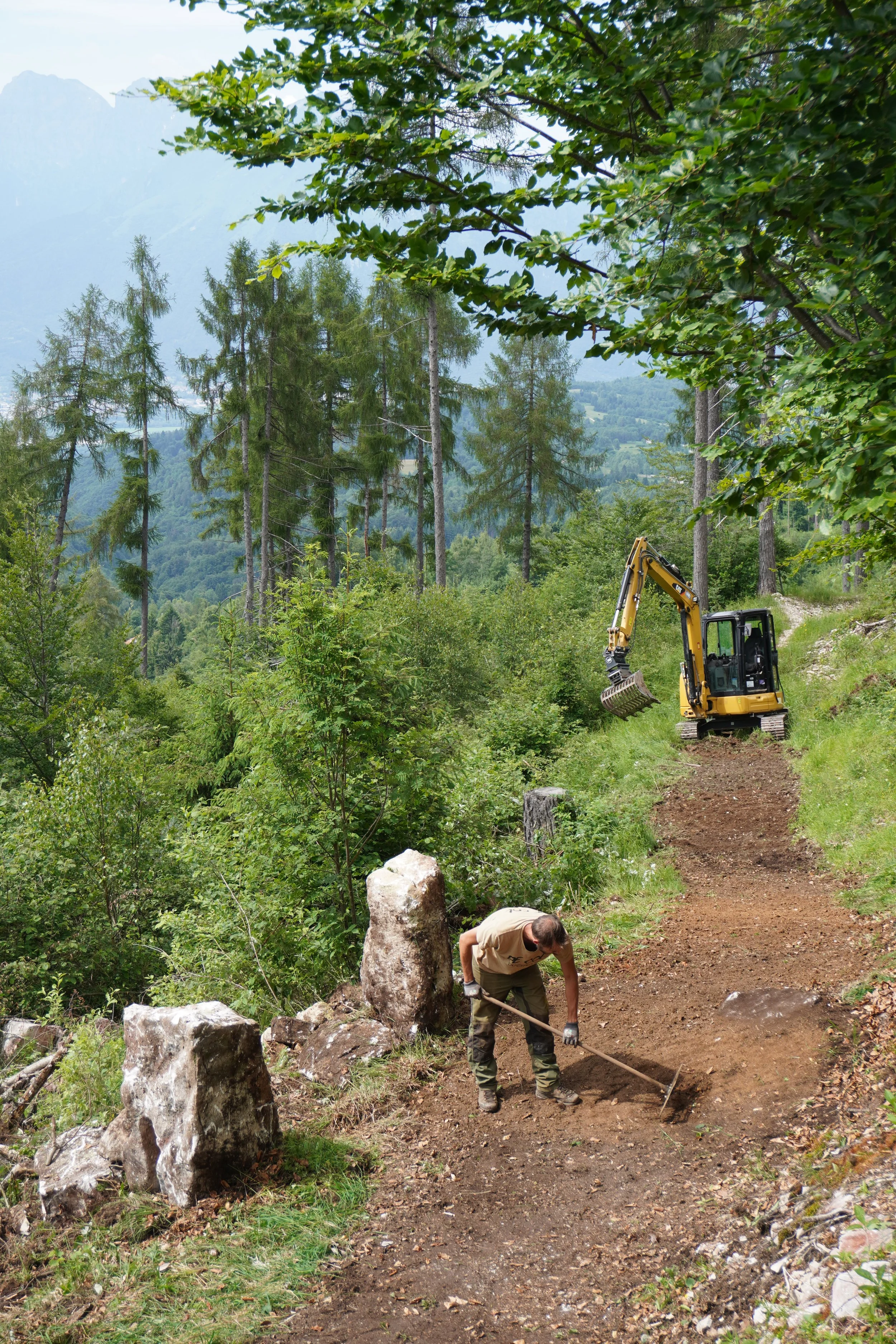 Un uomo sta lavorando alla manutenzione di un sentiero in una foresta, usando una pala, con un escavatore giallo sullo sfondo, tra alberi verdi e montagne in lontananza.