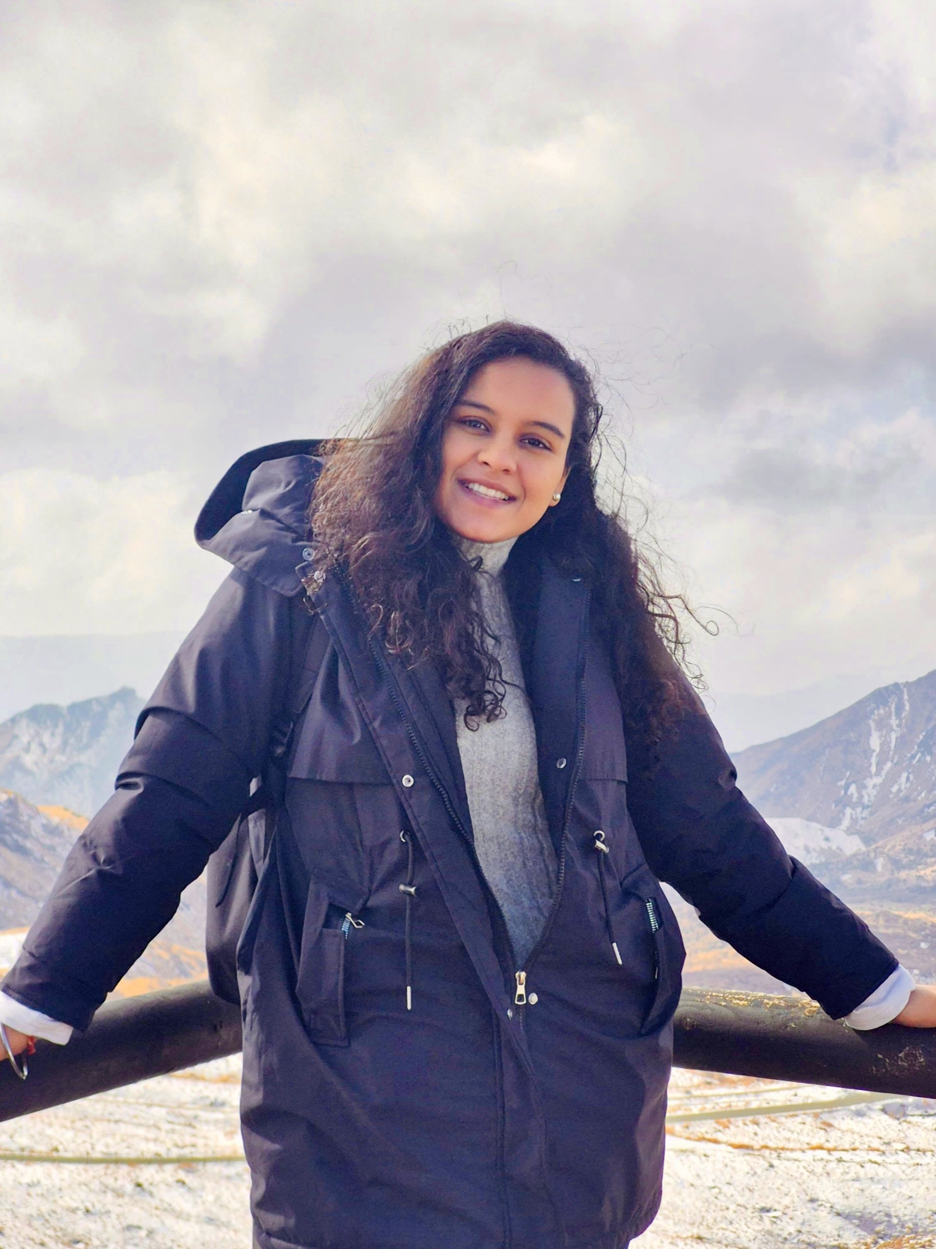 Young woman in a black winter jacket smiling outdoors with mountains and cloudy sky in the background.