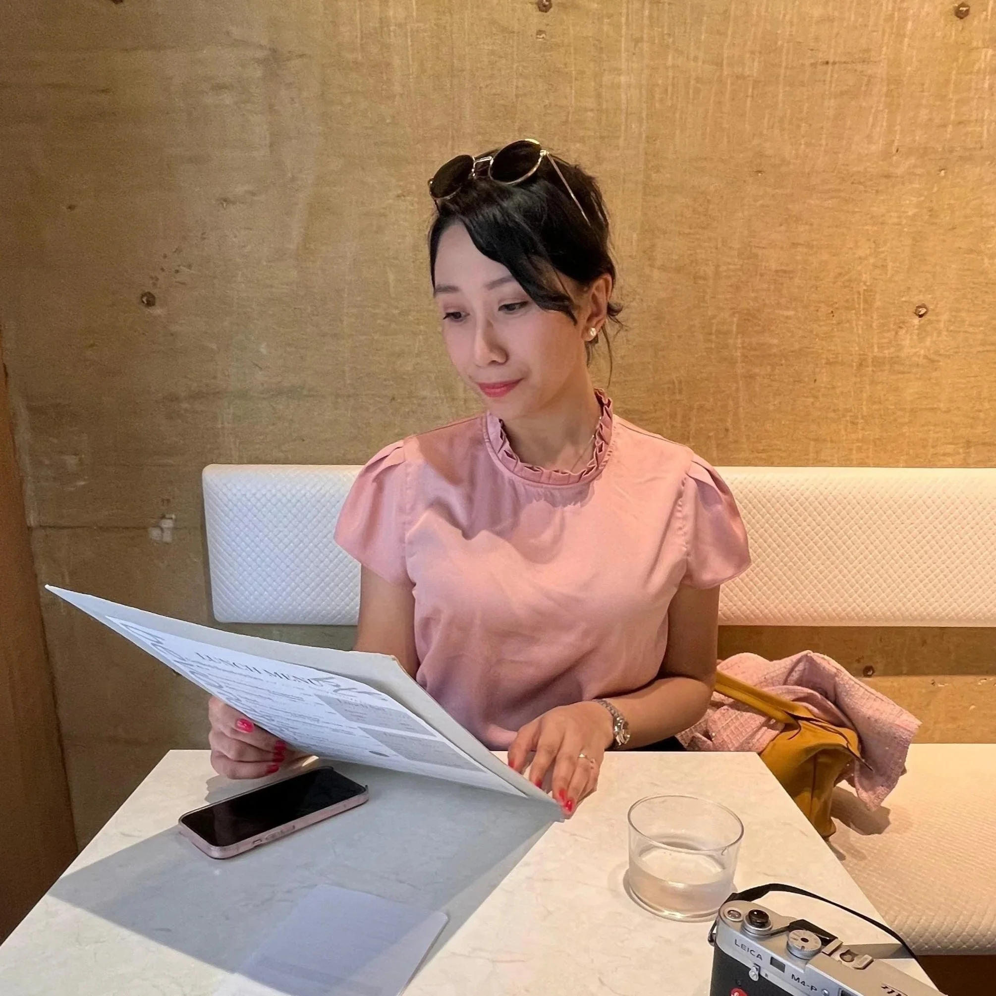 A woman sitting at a table in a restaurant, reading a menu. There is a camera, phone, and glasses on the table, along with a glass of water and a paper with the words "DIREK MEN" on it.