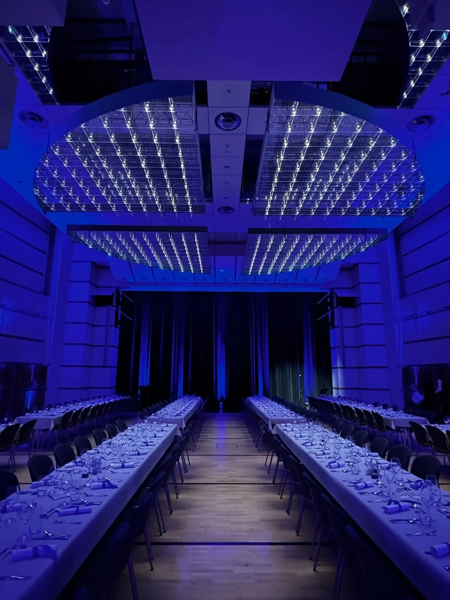 Ein festlich dekorierter Saal mit langen Tischen, gedeckt mit weißen Tischdecken, Gläser, Besteck und Servietten. Der Saal ist in Blau beleuchtet, mit einem großen, modernen Beleuchtungssystem an der Decke.