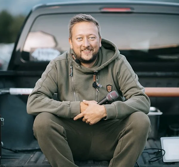Mann mit Bart, grauer Hoodie, sitzt vor einem schwarzen Fahrzeug und hält ein Mikrofon in der Hand.