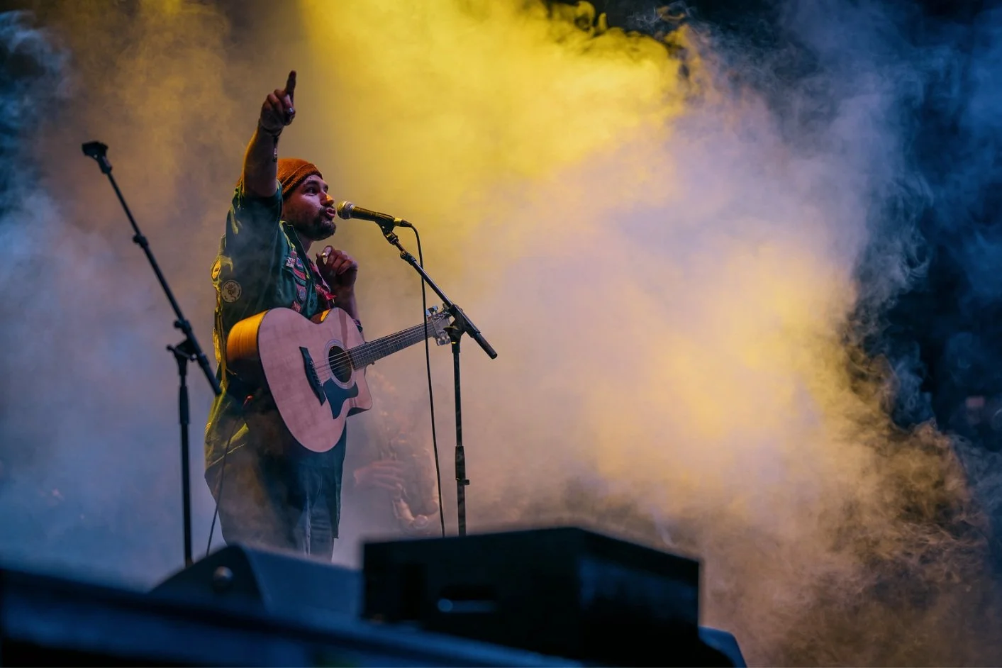 Musiker mit Gitarre und Beanie auf Bühne, umgeben von Rauch und bunter Beleuchtung, singt in Mikrofon.