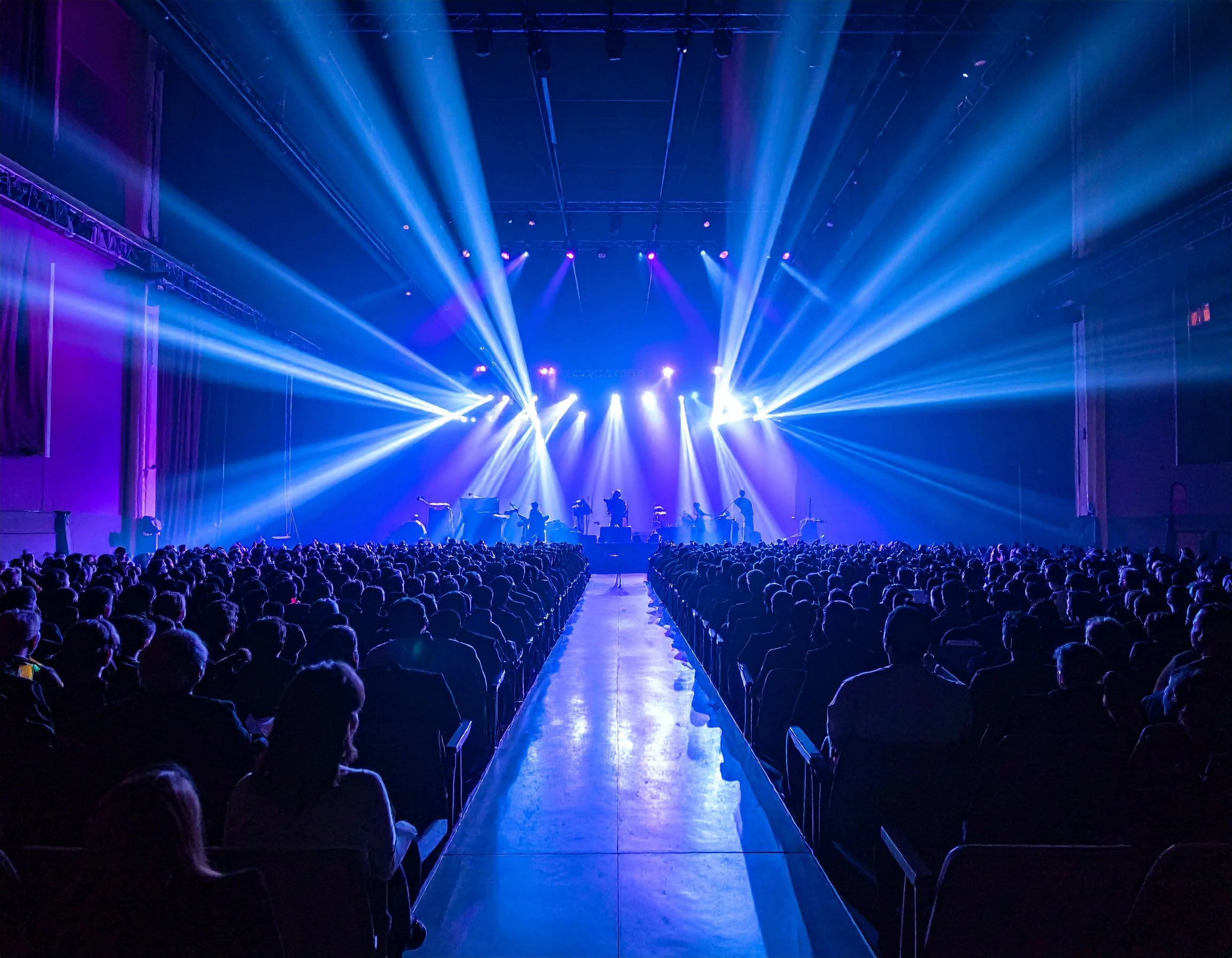 Bühne mit blauen Lichtstrahlen bei einem Konzert, mit Publikum im Saal.