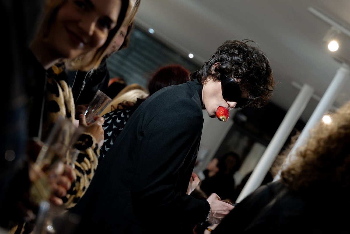 Photographie de reportage par Mélissa Pottier. Un homme surpris en train de manger une fraise lors d'un événement mode fashion week, atmosphère immersive captée avec spontanéité.