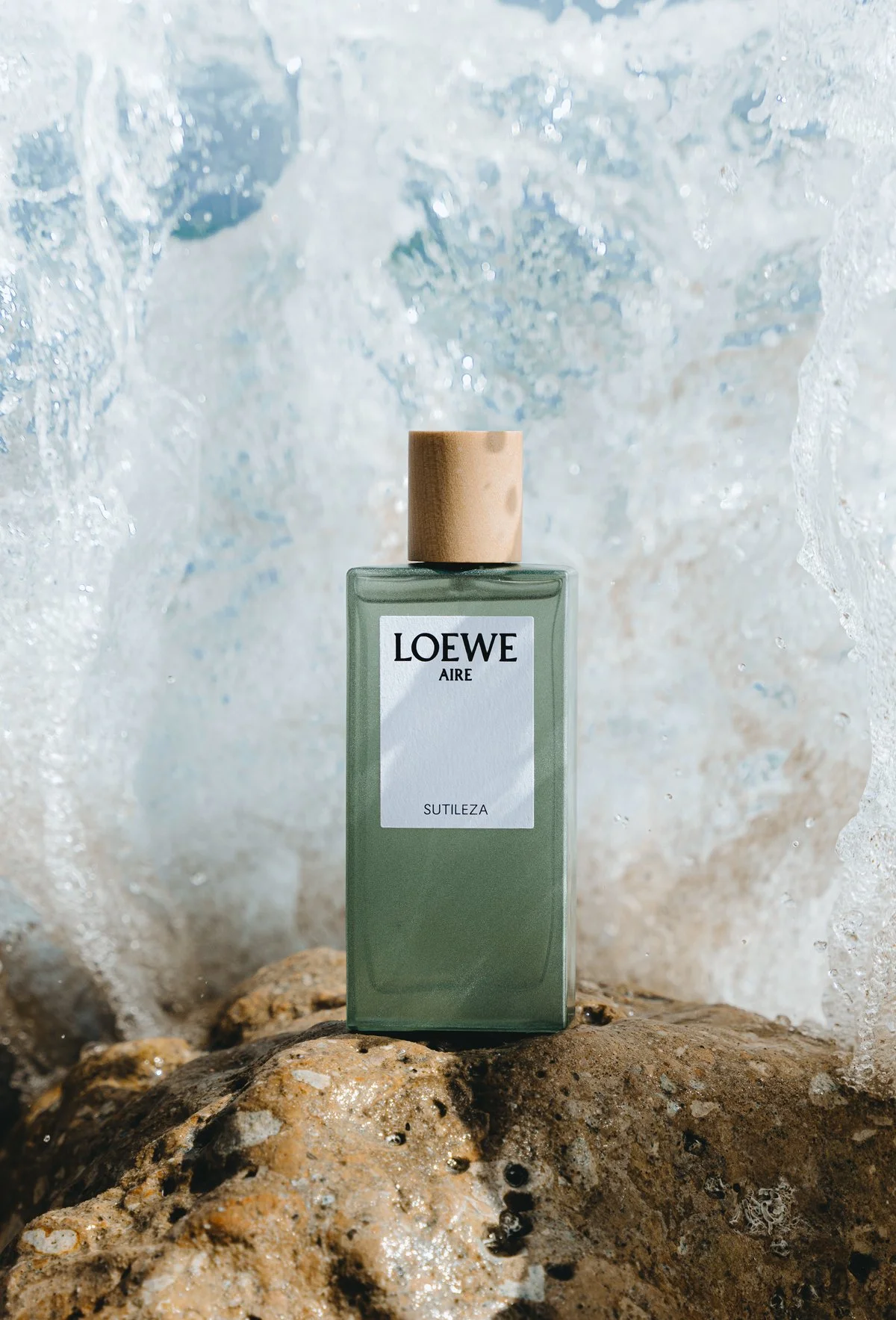 Une bouteille de parfum Loewe, repose sur un rocher et derrière une vague éclate