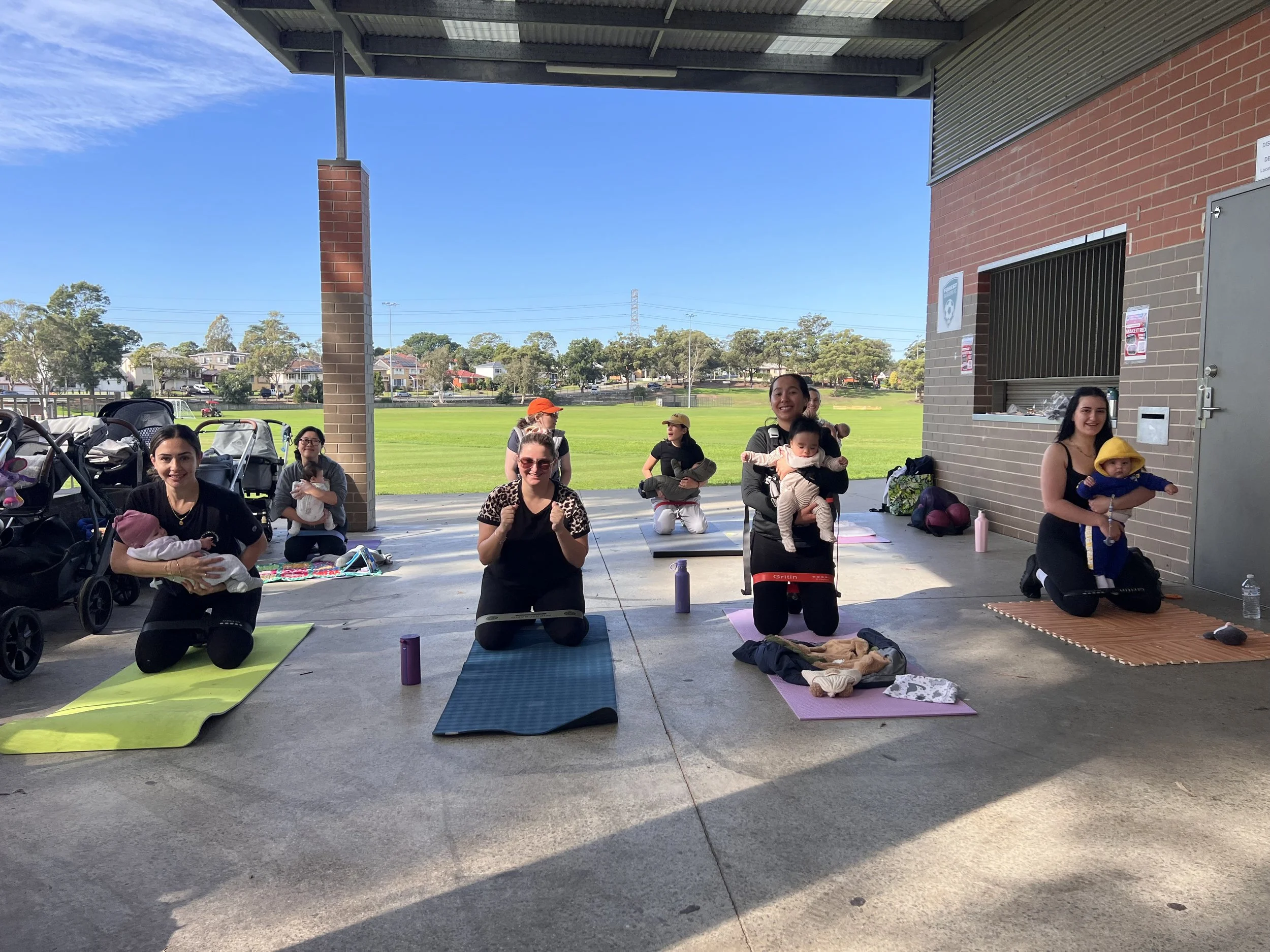Group of mums with babies participating in a Mums & Bubs fitness class outdoors under a covered area with a grassy field in the background.