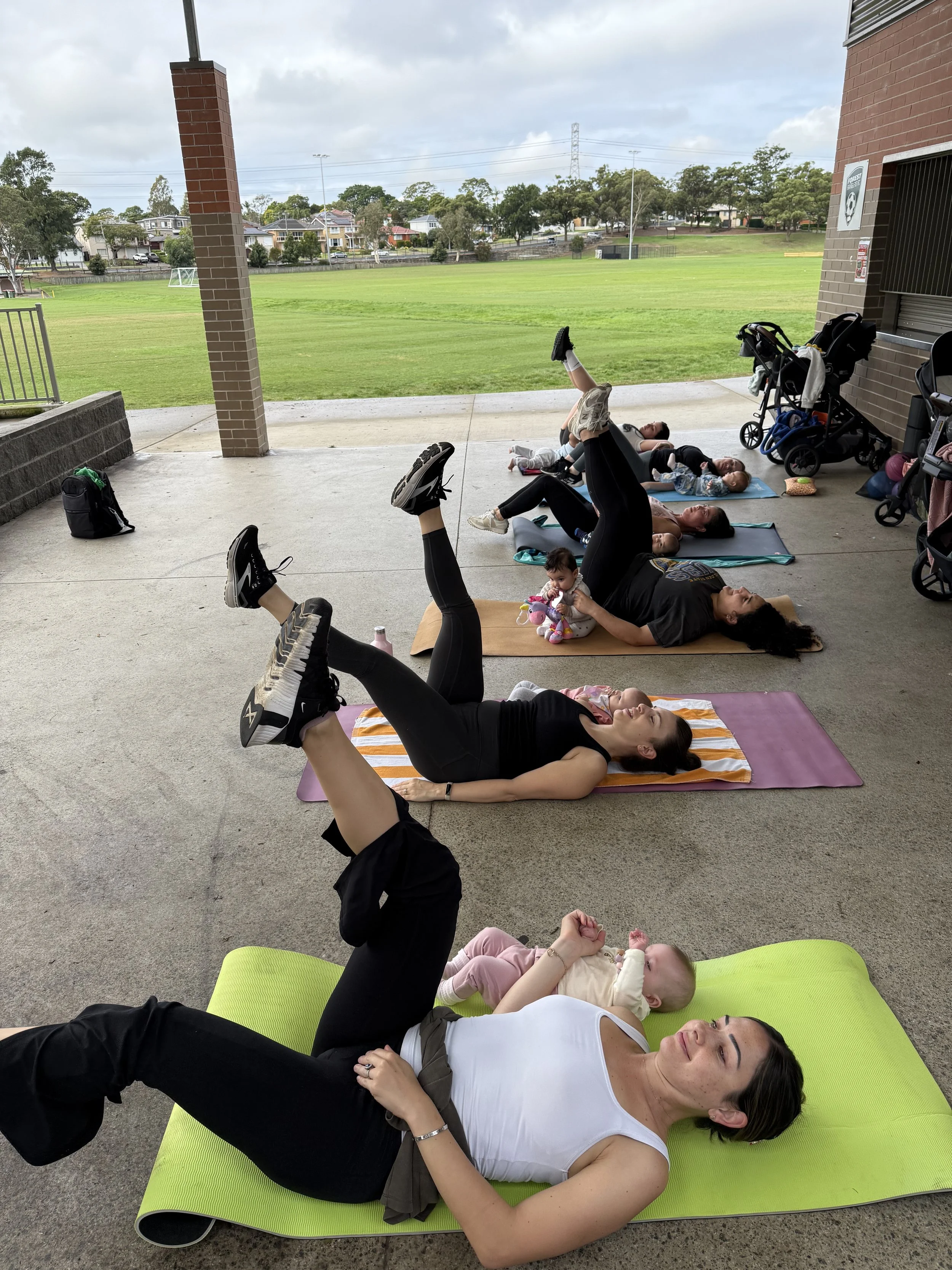 Group of mums and children participating in a baby yoga class, lying on exercise mats on an outdoor covered pavilion, with a grassy field and houses in the background.