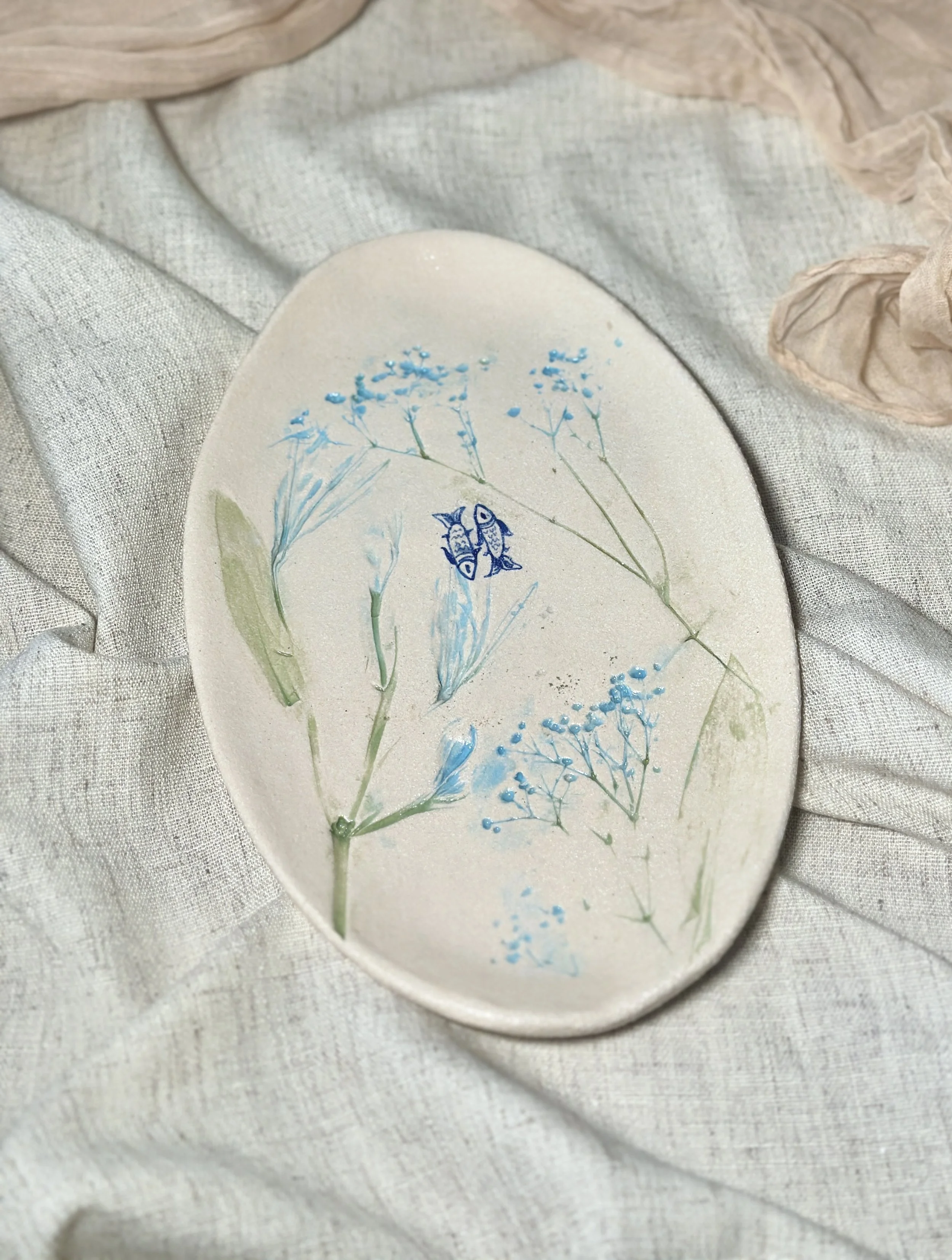 Decorative oval dish with pressed blue flowers and a tiny illustration of a butterfly, resting on light-colored fabric.