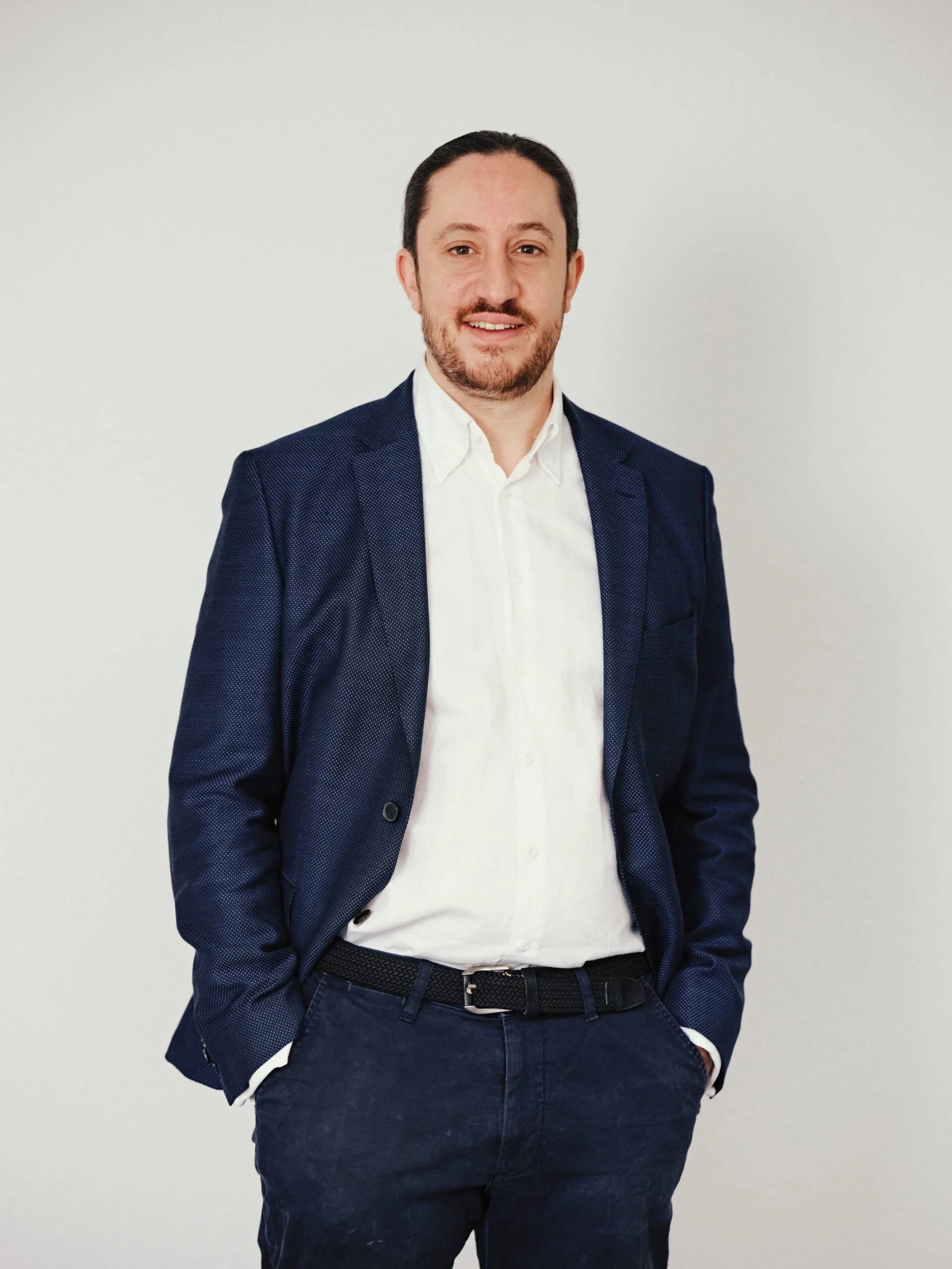 Ein Mann trägt einen dunklen Anzug und ein weißes Hemd, steht vor einem weißen Hintergrund und lächelt.
