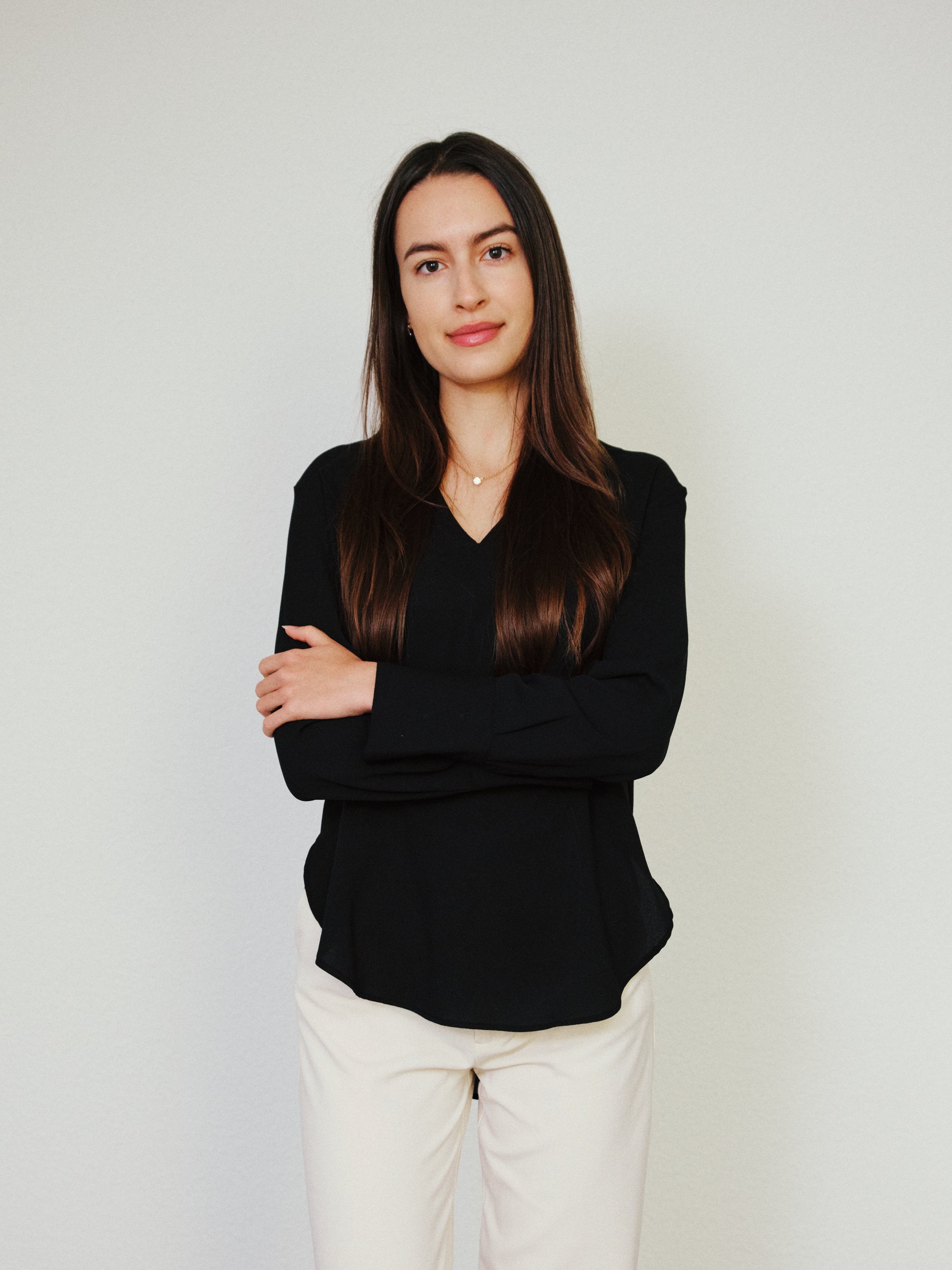 Eine junge Frau steht vor einem neutralen weißen Hintergrund, trägt eine schwarze Bluse und weiße Hosen, hat braune Haare und ein Lächeln.