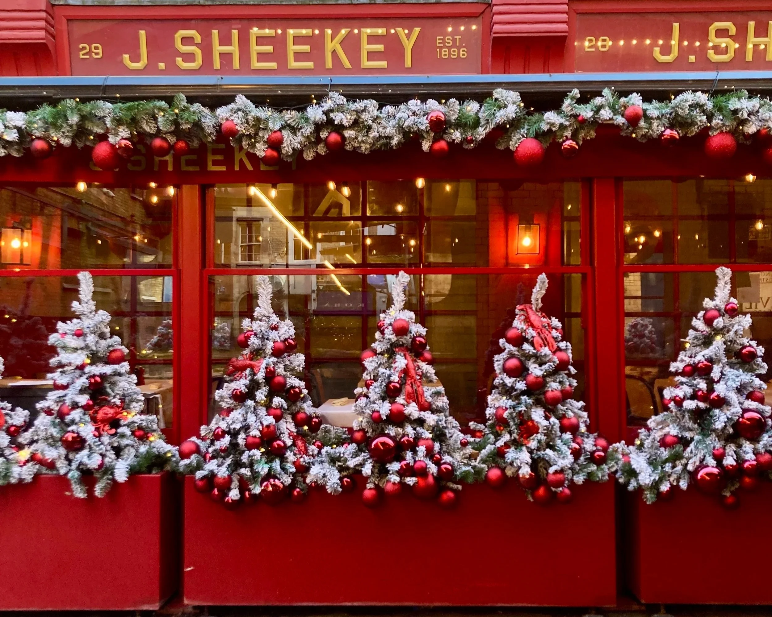 Festively decorated storefront with large Christmas wreaths and garlands featuring red, green, gold, and white ornaments, peppermint candies, and red ribbons, flanked by potted plants and outdoor benches.