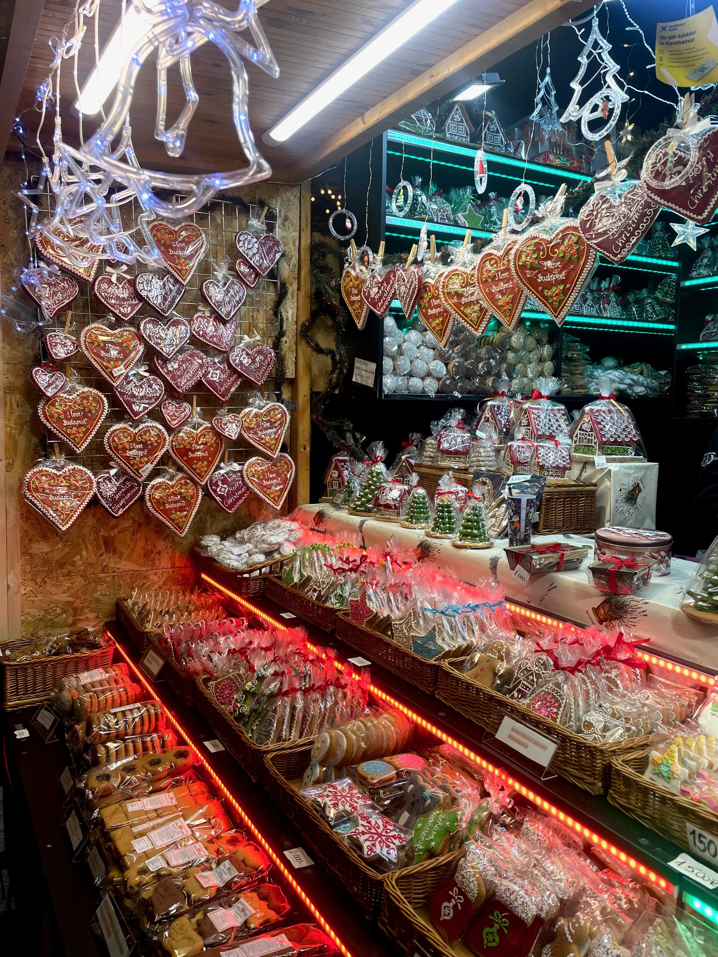 Festively decorated storefront with large Christmas wreaths and garlands featuring red, green, gold, and white ornaments, peppermint candies, and red ribbons, flanked by potted plants and outdoor benches.