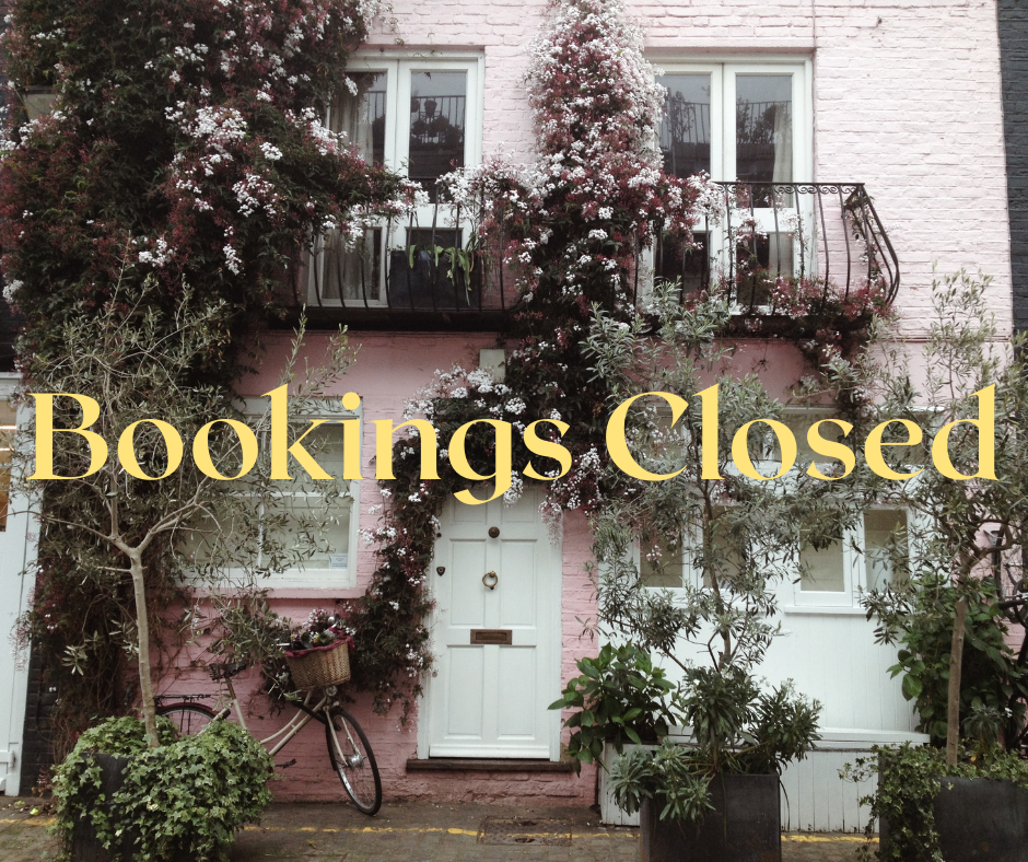 Pink brick house with white door and multiple window, decorated with pink and white flowering plants, a bicycle with a basket leaning against the wall, and various potted plants in front.