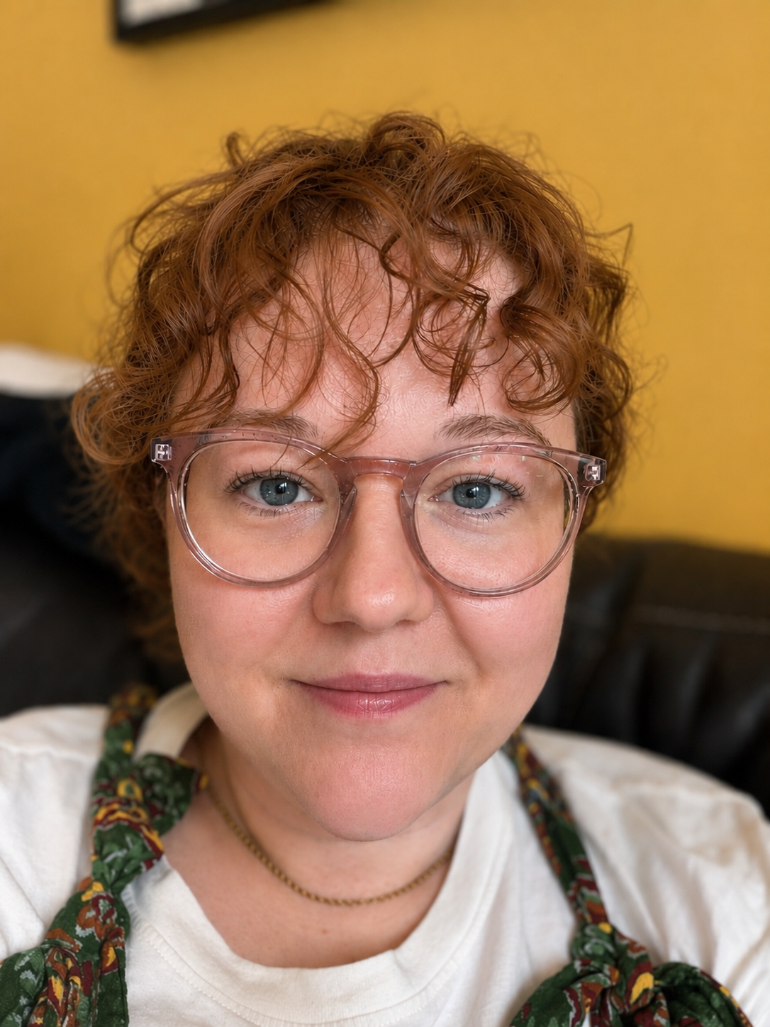 A woman with short, curly red hair and glasses smiling at the camera, wearing a white shirt and a floral scarf, against a yellow background.