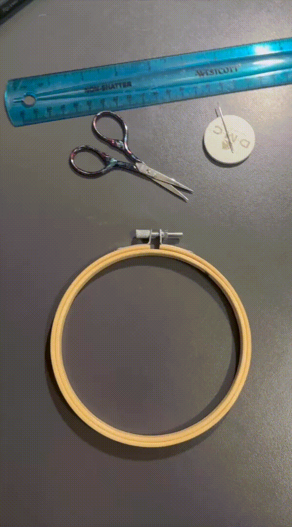 Materiales de costura sobre una mesa, incluyendo una regla, tijeras, un reloj pequeño y un aro de bordado con bastidor.