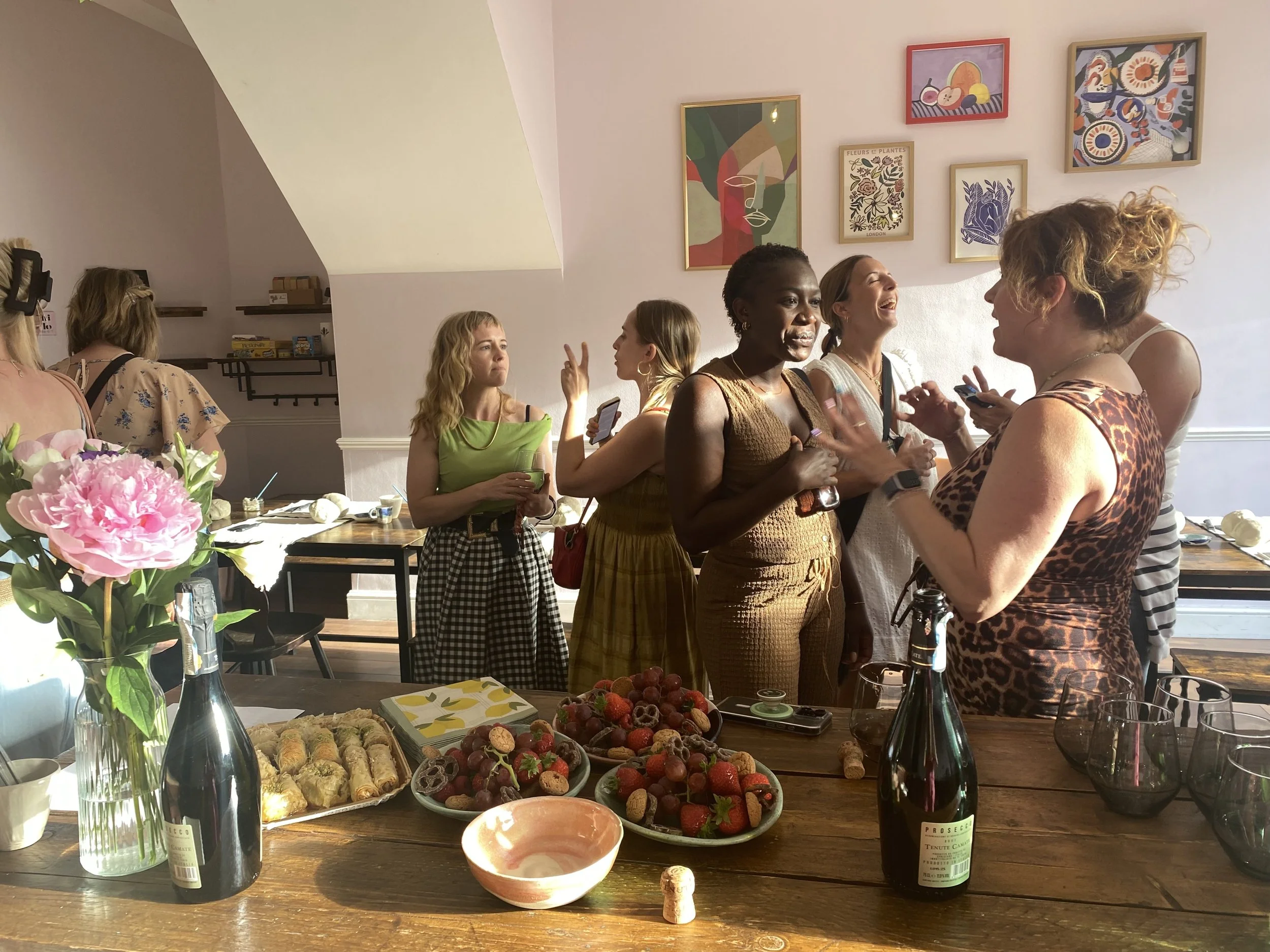 Group of women socializing and laughing around a table with an array of desserts, wine, and flowers, in a brightly decorated room.