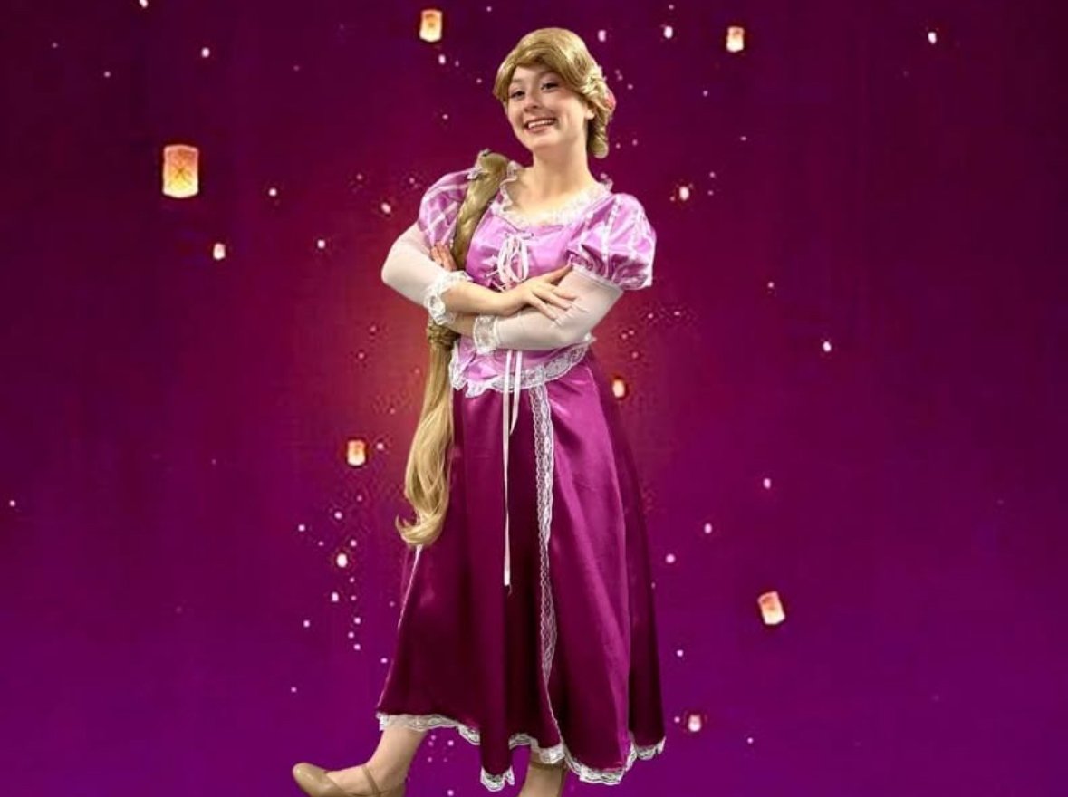 A woman dressed as Rapunzel from Disney's Tangled, wearing a pink and purple dress, standing in front of a purple background with floating lanterns. Lake Buena Vista