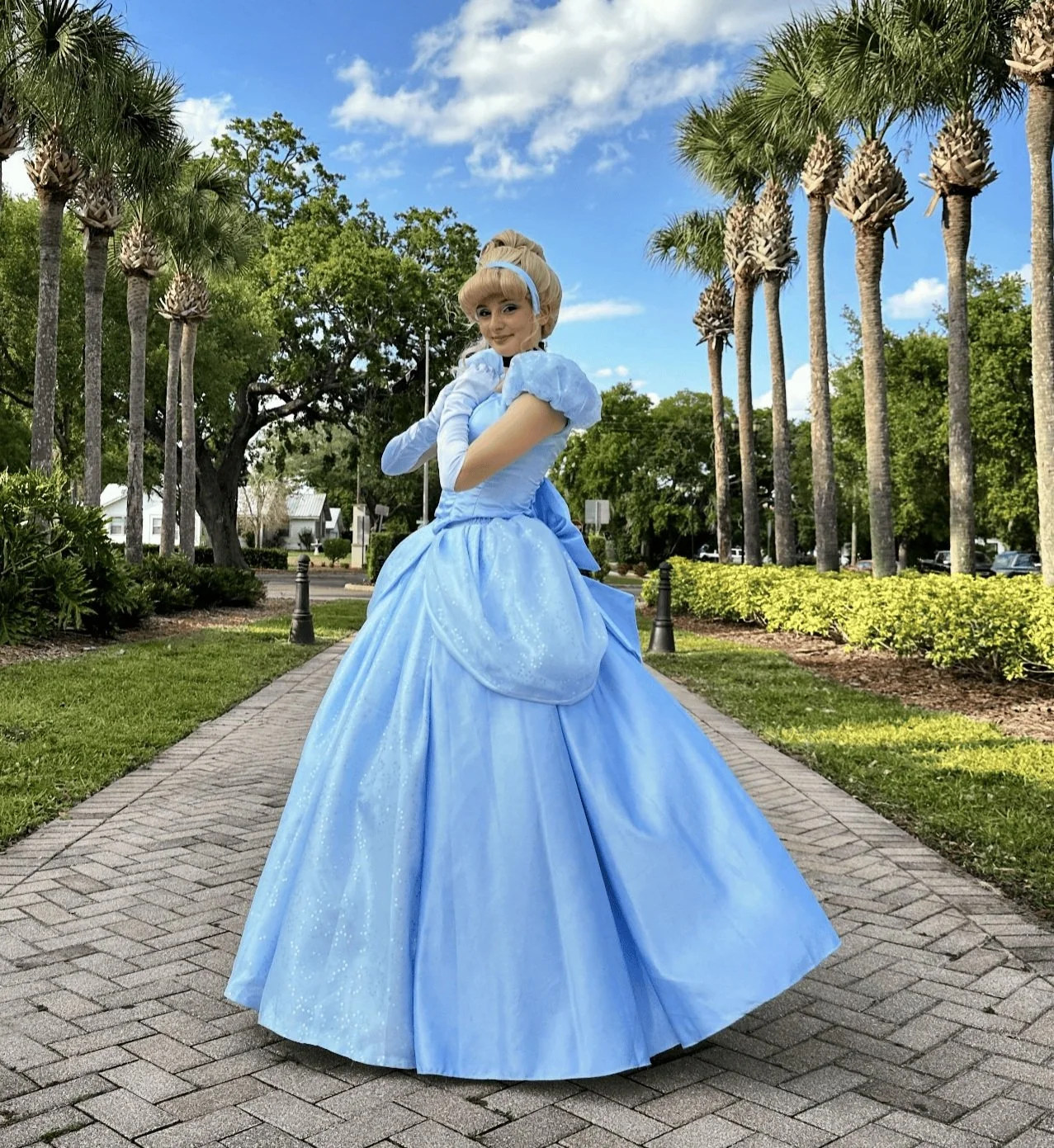 A woman dressed as Cinderella in a light blue gown standing on a brick pathway in a park, with palm trees, greenery, and a blue sky with white clouds in the background. Polk County