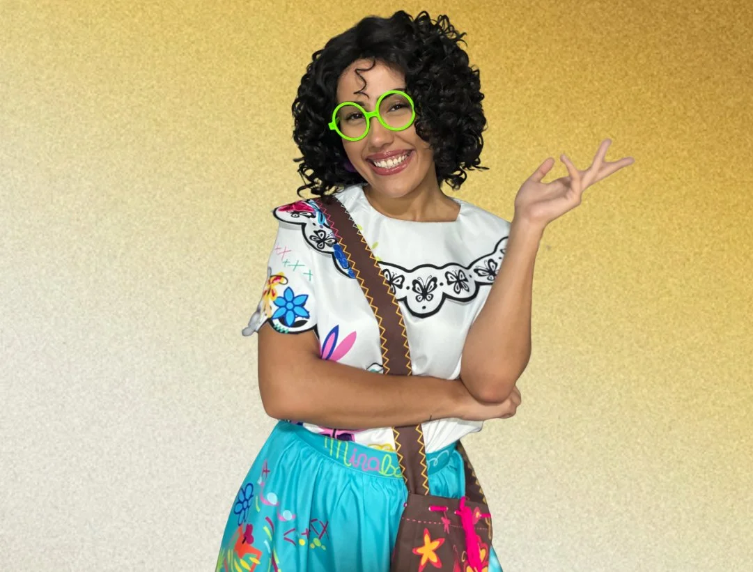 A woman dressed in a colorful outfit with curly hair and green glasses, smiling confidently against a gold gradient background. Mirabel From Encanto Princess Party Characters Orlando