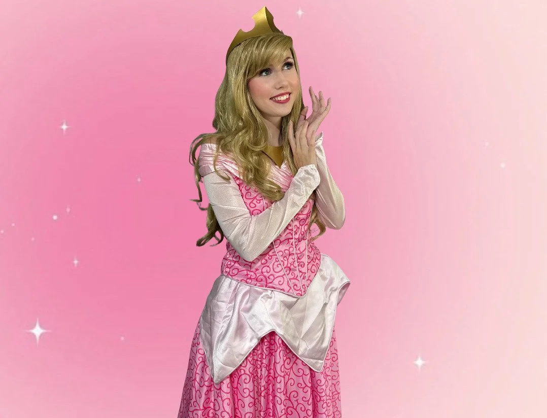 A woman dressed as a princess with a pink and white dress, a gold crown, and long blonde hair, standing against a pink background with white sparkles.