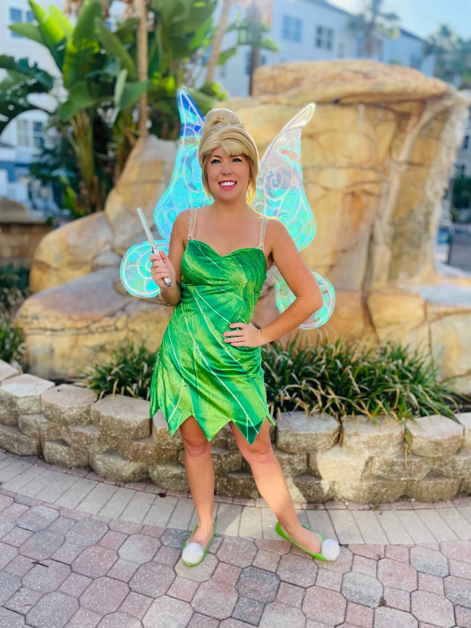 Woman dressed as Tinker Bell in a green leaf costume, fairy wings, and holding a magic wand, standing in front of a decorative fountain with plants.