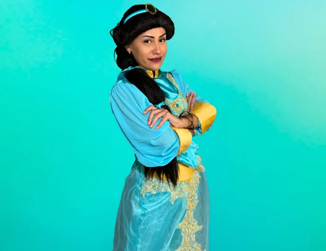 A woman dressed as Princess Jasmine from Aladdin, wearing a blue and gold costume with long black hair, standing in front of a gradient blue-green background.