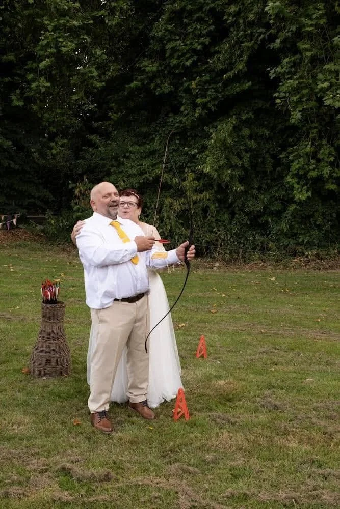 Bride and groom laughing as they do archery together in their wedding dress and suit