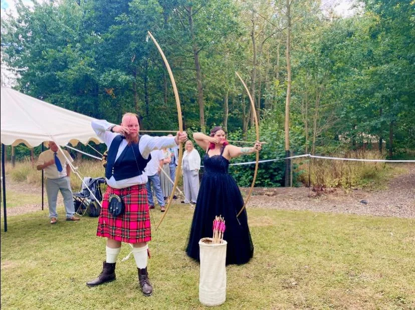 Bride in black wedding dress and groom in kilt shooting longbows in a wood