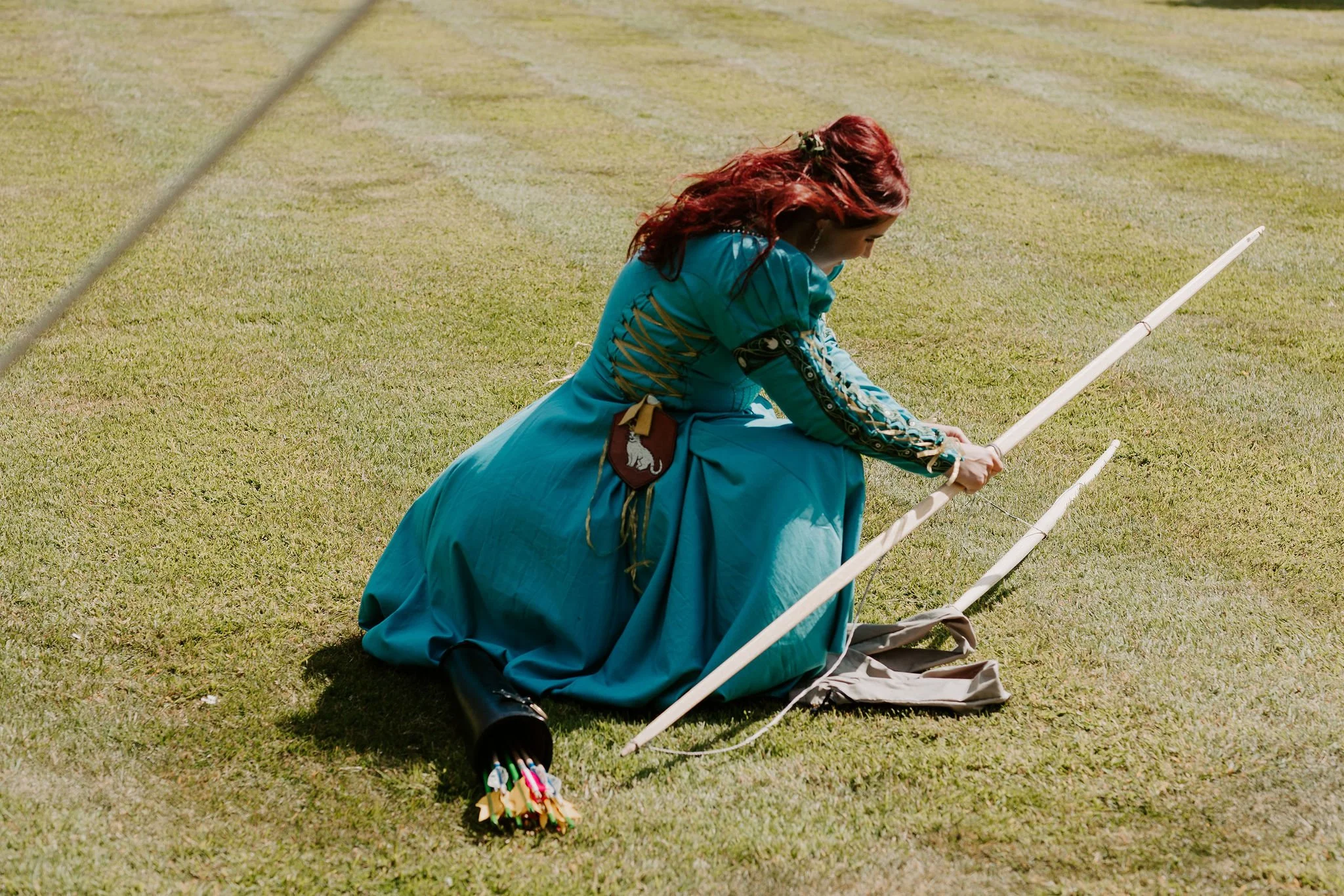 Woman with flowing red hair and medieval-style teal dress stringing a longbow on a lawn