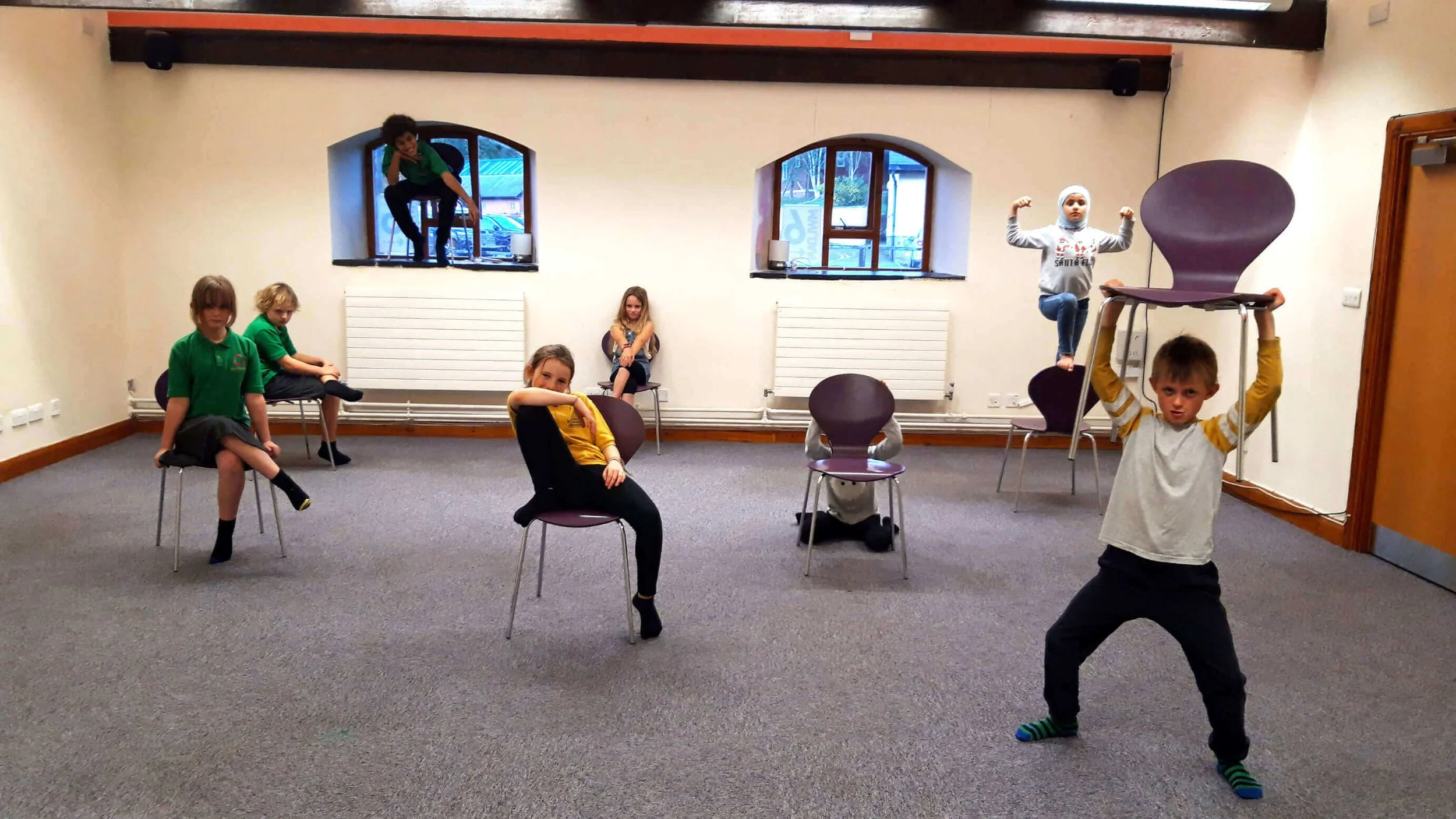 Children in a room playing and posing, some sitting on chairs, one girl in a yellow shirt and another girl in a hijab and blue jeans, a boy holding a chair overhead, and a girl perched on a high chair near the window.