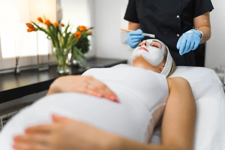 A woman lying on a treatment bed with a facial mask being applied by a professional wearing blue gloves, in a spa or clinic setting.
