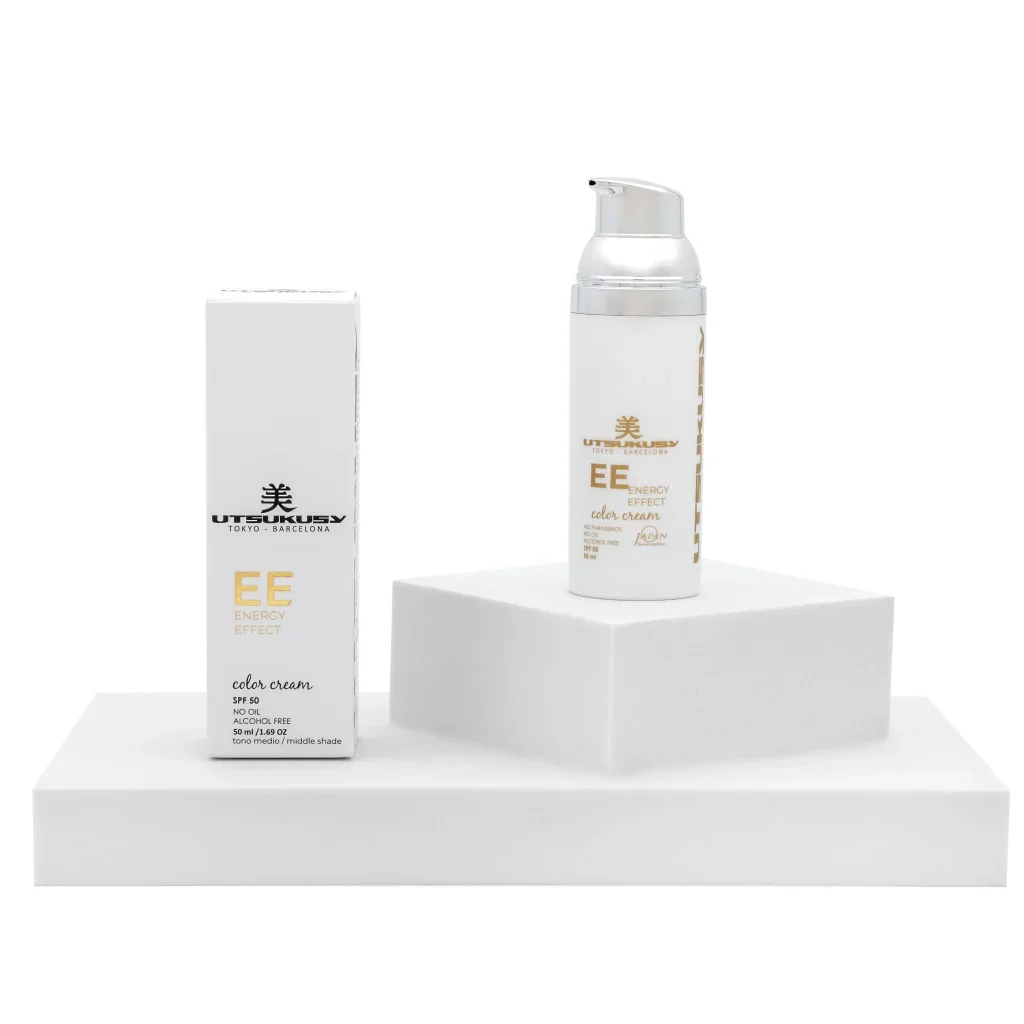 Dos envases de crema de color con etiqueta blanca y dorada, uno en una caja y otro en un soporte elevado, en fondo blanco.